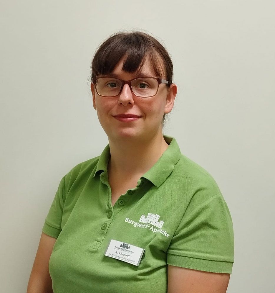 Frau mit Brille, grünem Poloshirt mit Namensschild, lächelnd im Innenbereich.