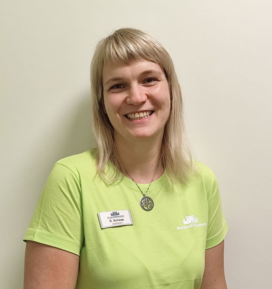Frau mit blondem Haar, lächelnd, mit grünem T-Shirt und Namensschild, vor einer weißen Wand.