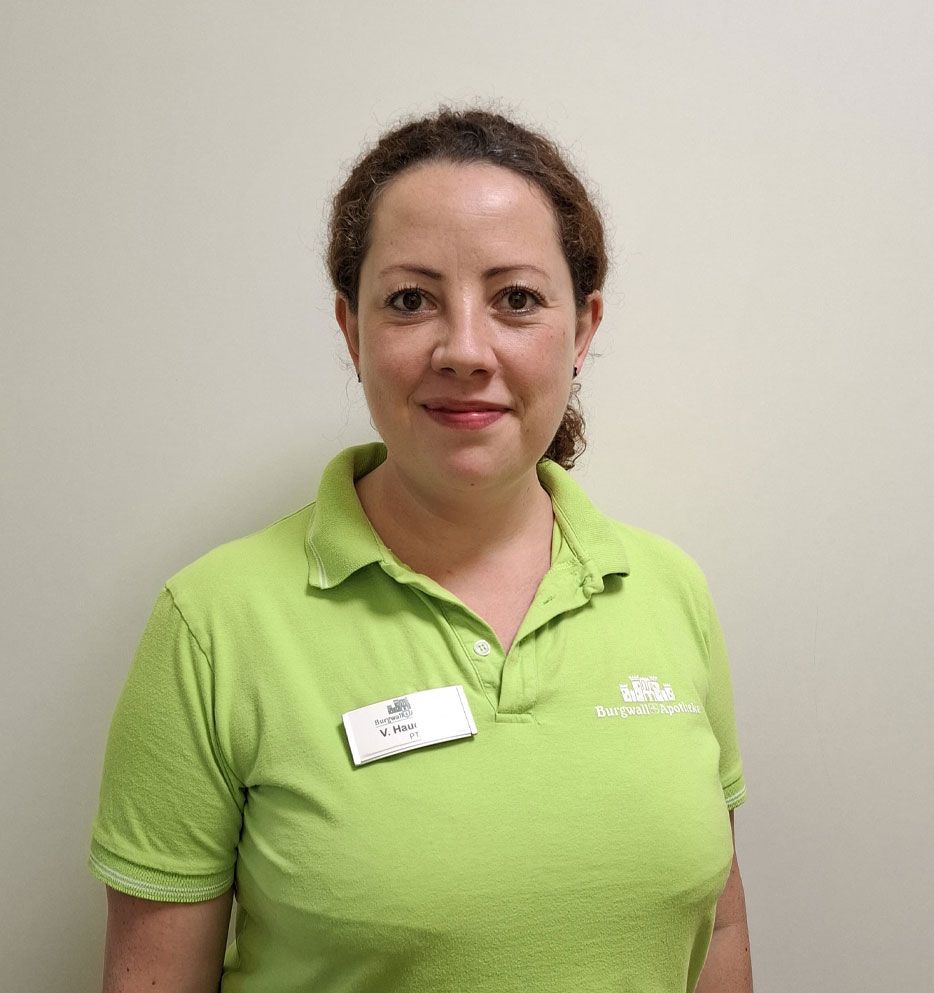 Frau mit lockigem Haar, grünem Poloshirt, lächelnd, mit Namensschild, vor einer schlichten Wand.