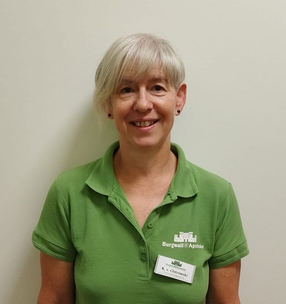 Frau mit kurzen grauen Haaren, die ein grünes Poloshirt mit Namensschild trägt und vor einer schlichten Wand lächelt.