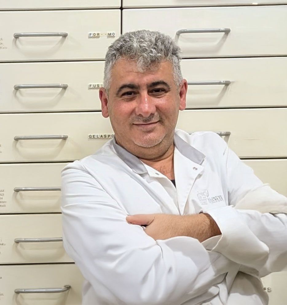 Ein Mann im weißen Laborkittel lächelt mit verschränkten Armen vor Apothekenschubladen.