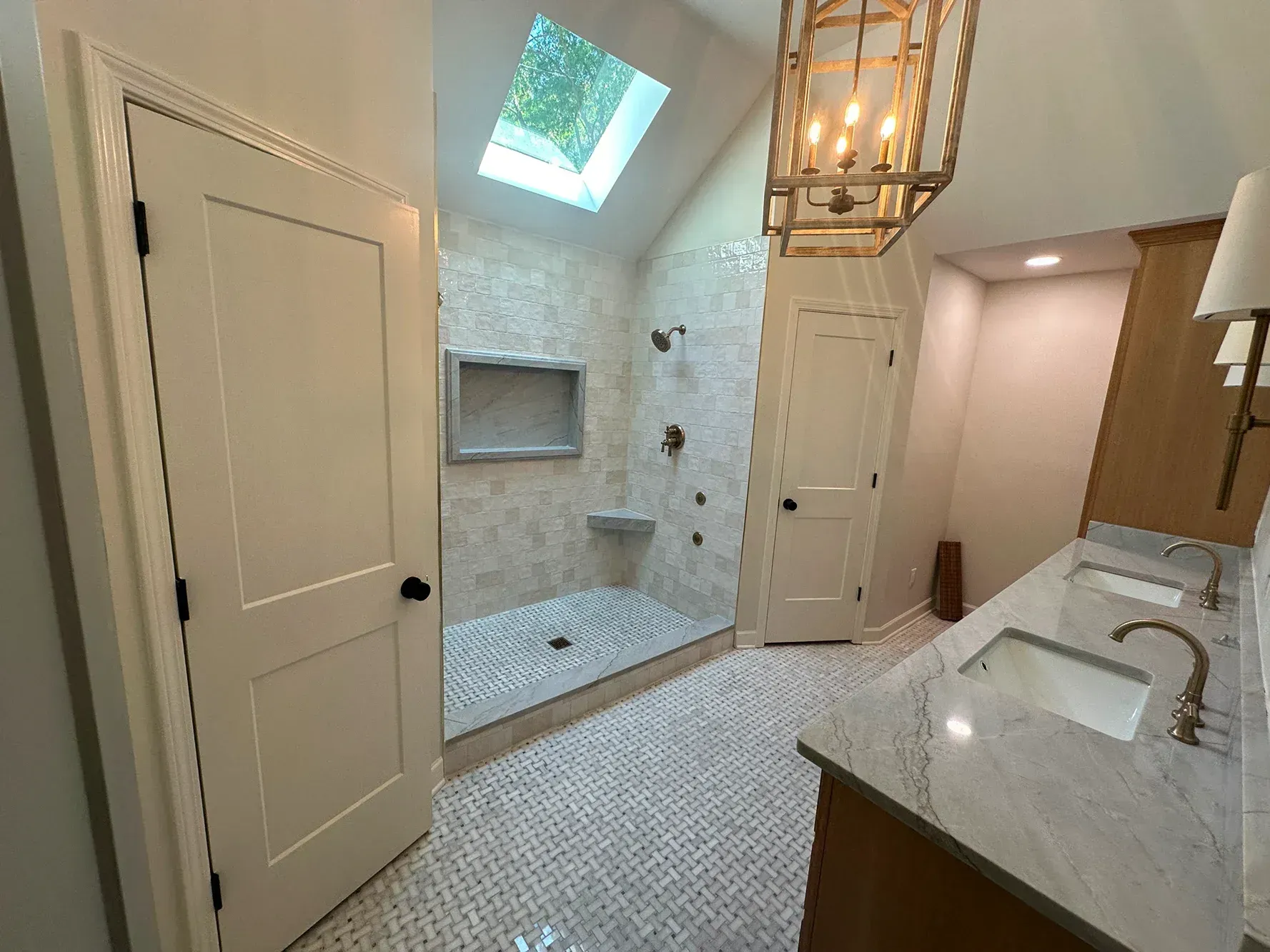 Bathroom with shower, vanity, and skylight. White and beige tiles, light wood cabinets, and brass chandelier.