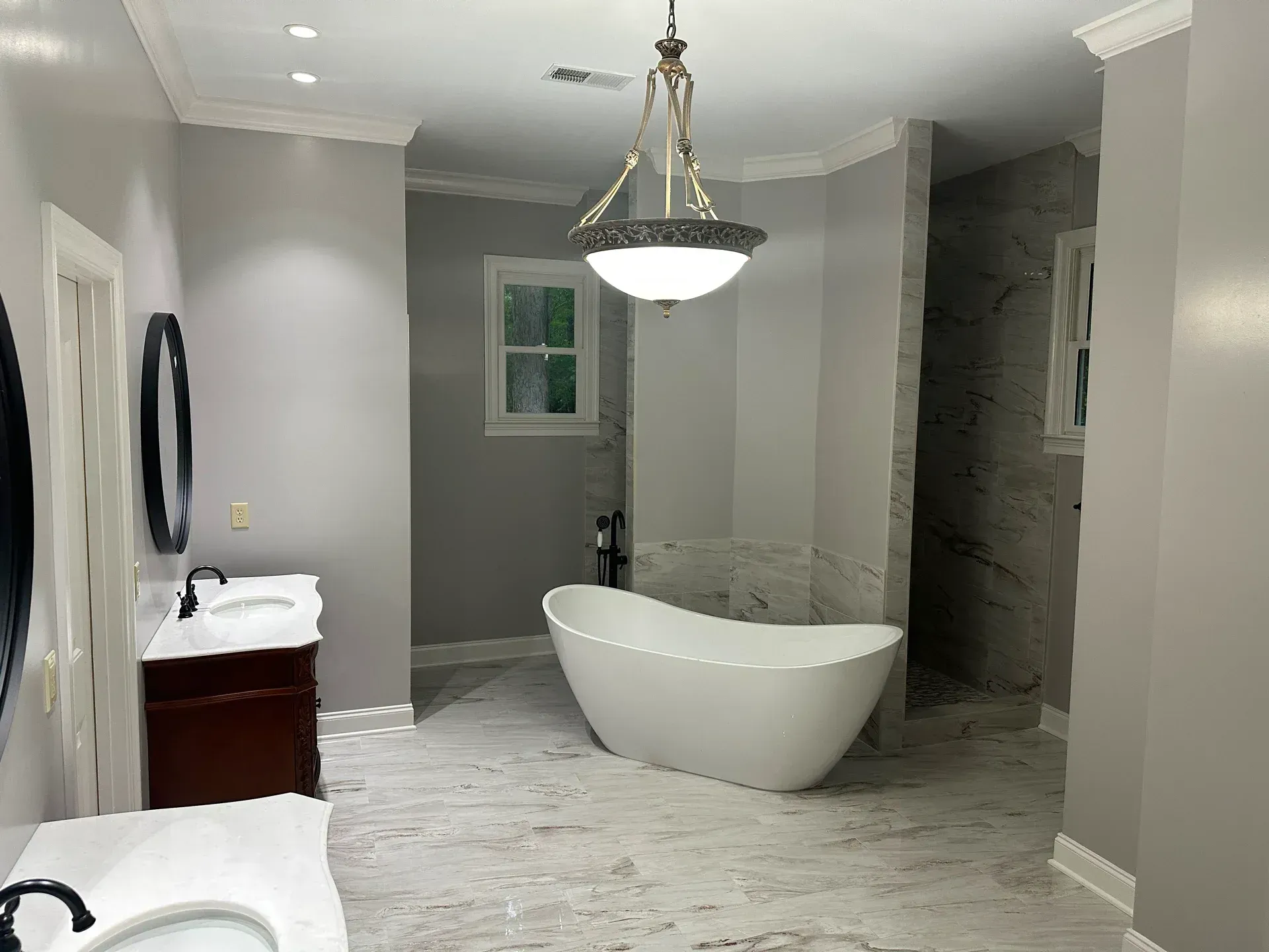 Spacious, modern bathroom with a white tub, two vanities, and a walk-in shower. Gray walls and marble flooring.