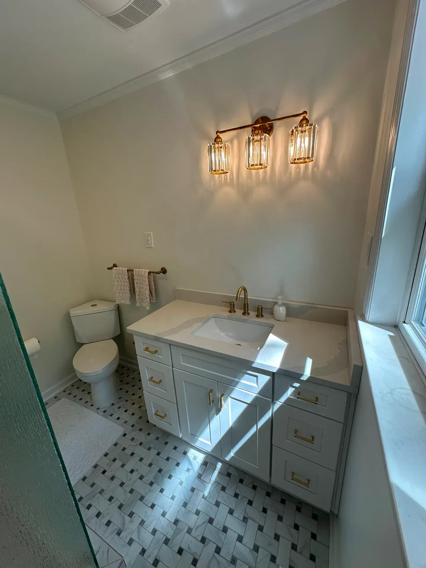 Bathroom with gray vanity, gold fixtures, and a three-light sconce. White toilet and patterned floor.