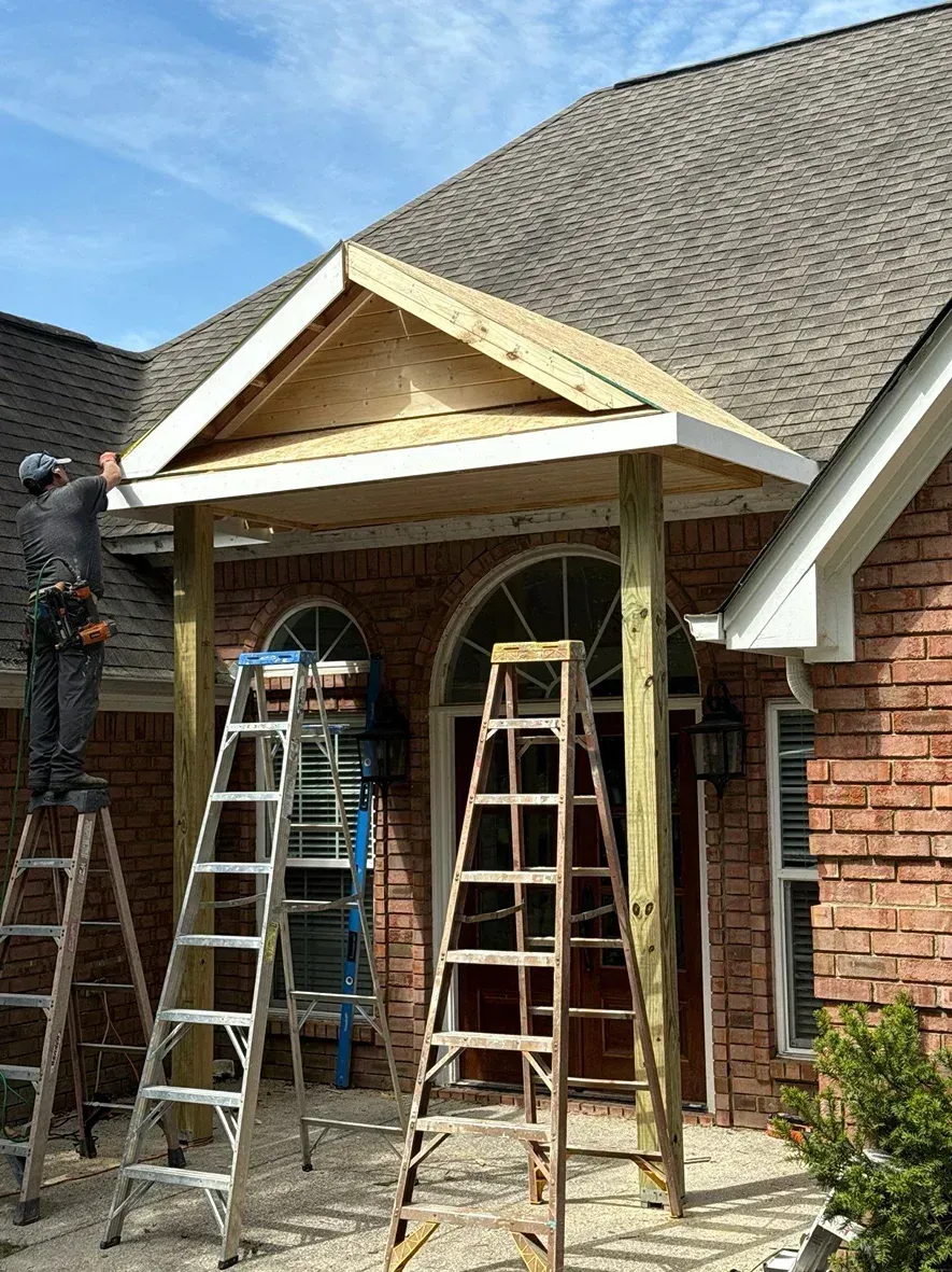 Construction of a porch roof on a brick house; workers on ladders, wood framing, and blue sky.