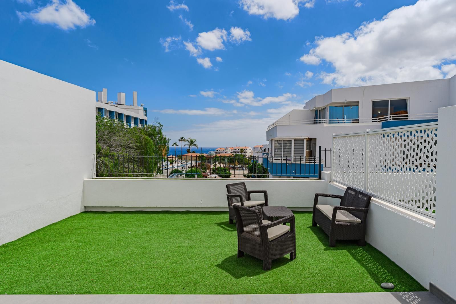 Pequeña terraza en la azotea con césped verde, sillas de patio negras y paredes blancas bajo un cielo azul.