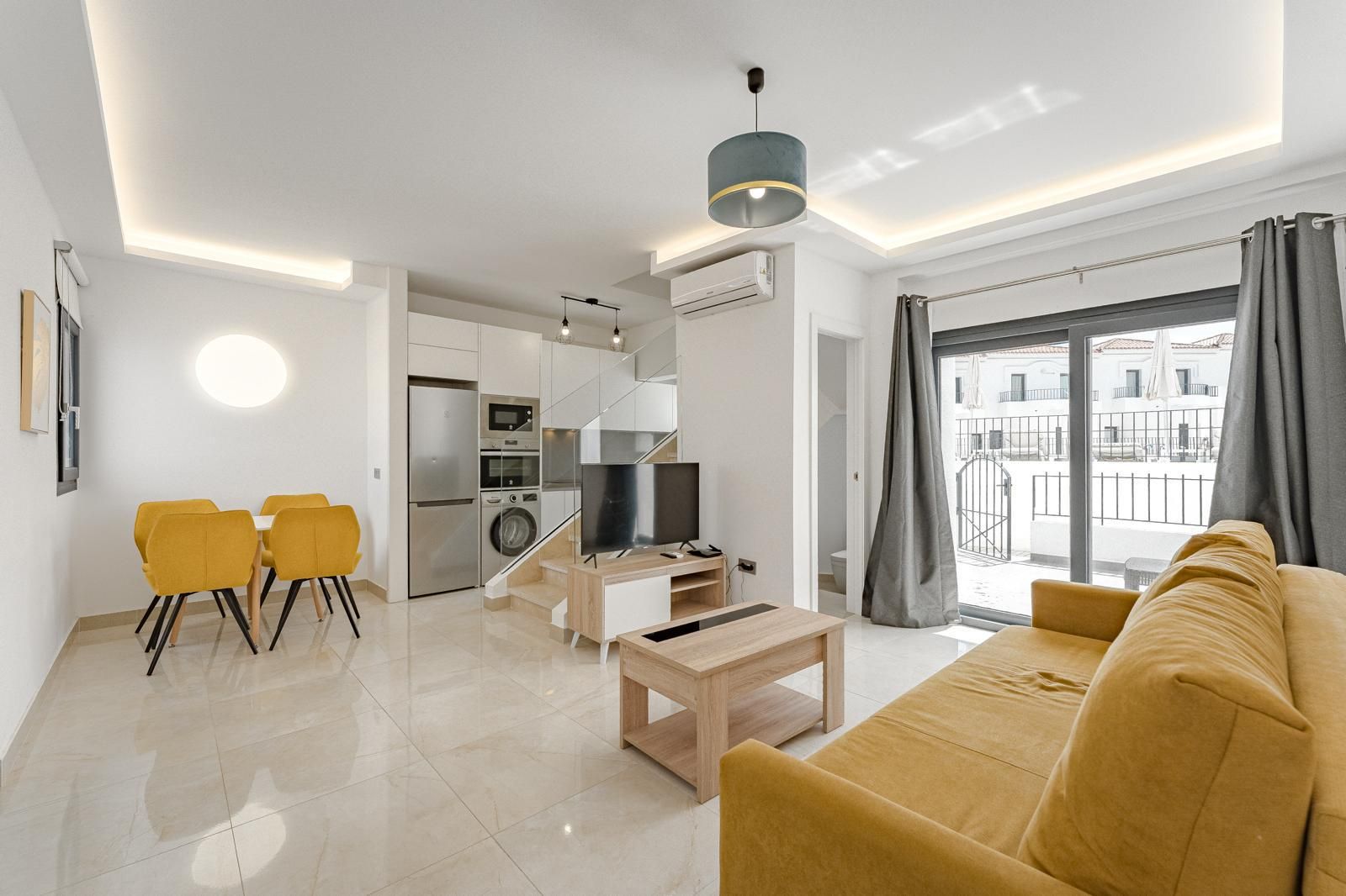 Sala moderna y luminosa con sofá mostaza, comedor, TV y grandes puertas al balcón con cortinas grises.