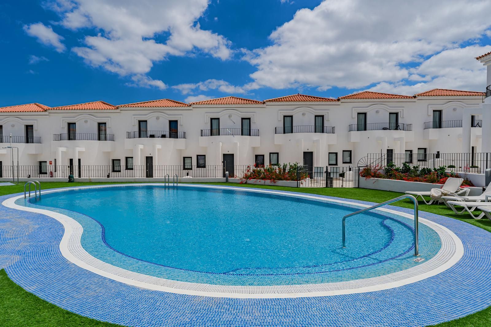 Piscina curva de color turquesa frente a villas blancas del complejo bajo un cielo azul.