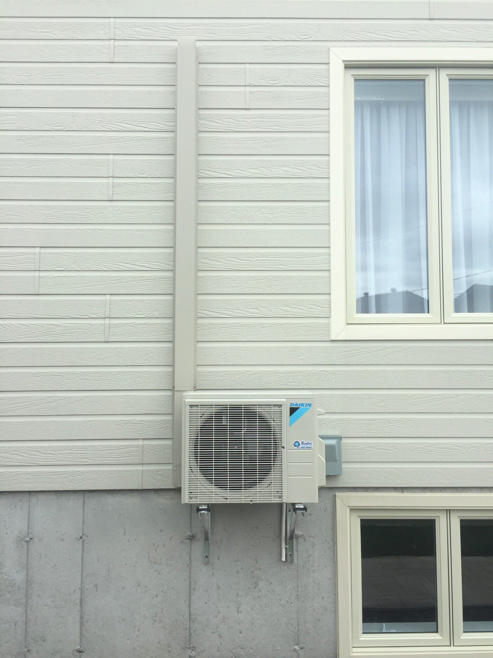 Unité de climatisation extérieure montée sur un mur à revêtement clair, à côté d'une fenêtre.