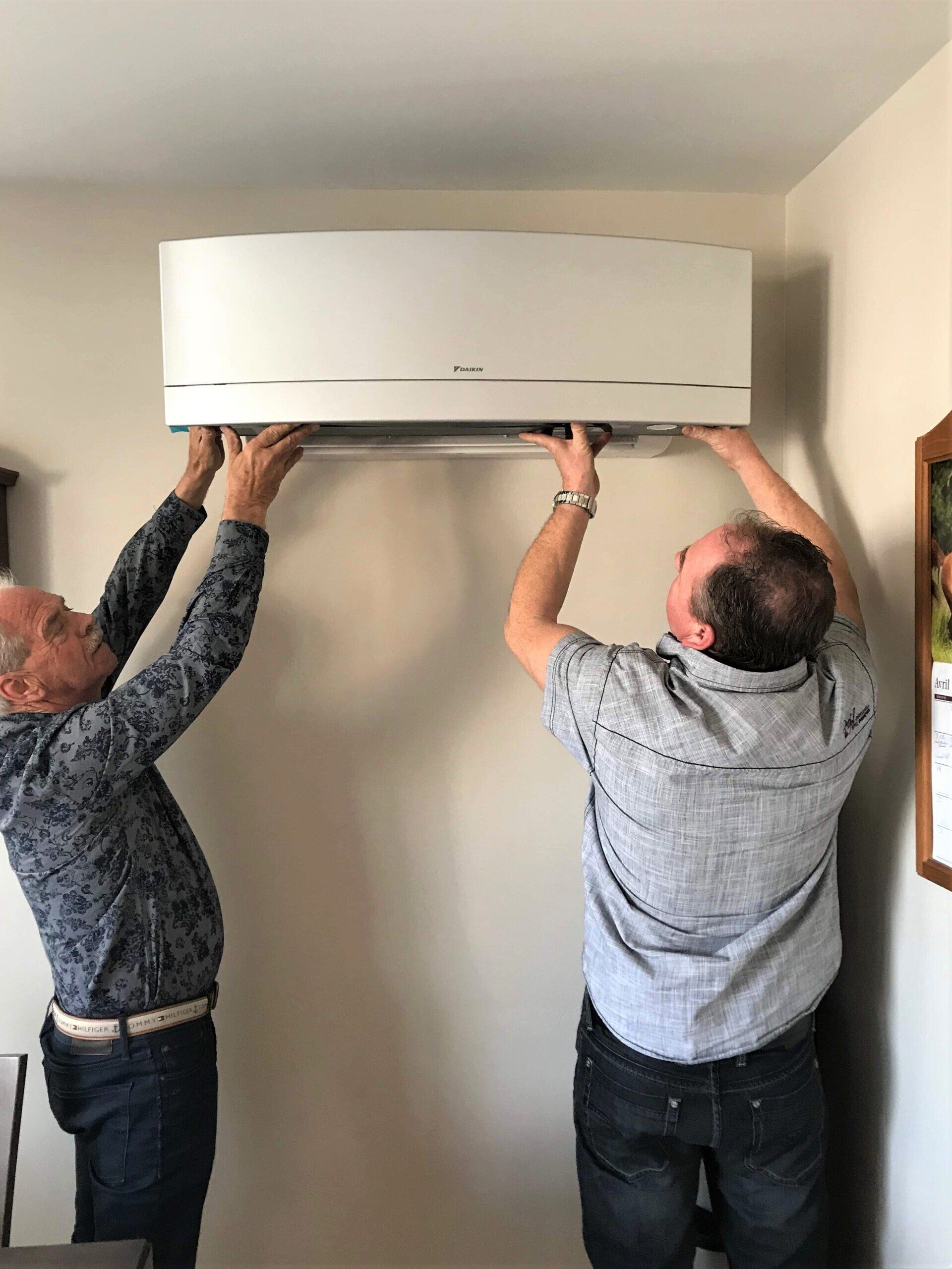 Deux hommes installent un climatiseur blanc sur un mur beige à l'intérieur.