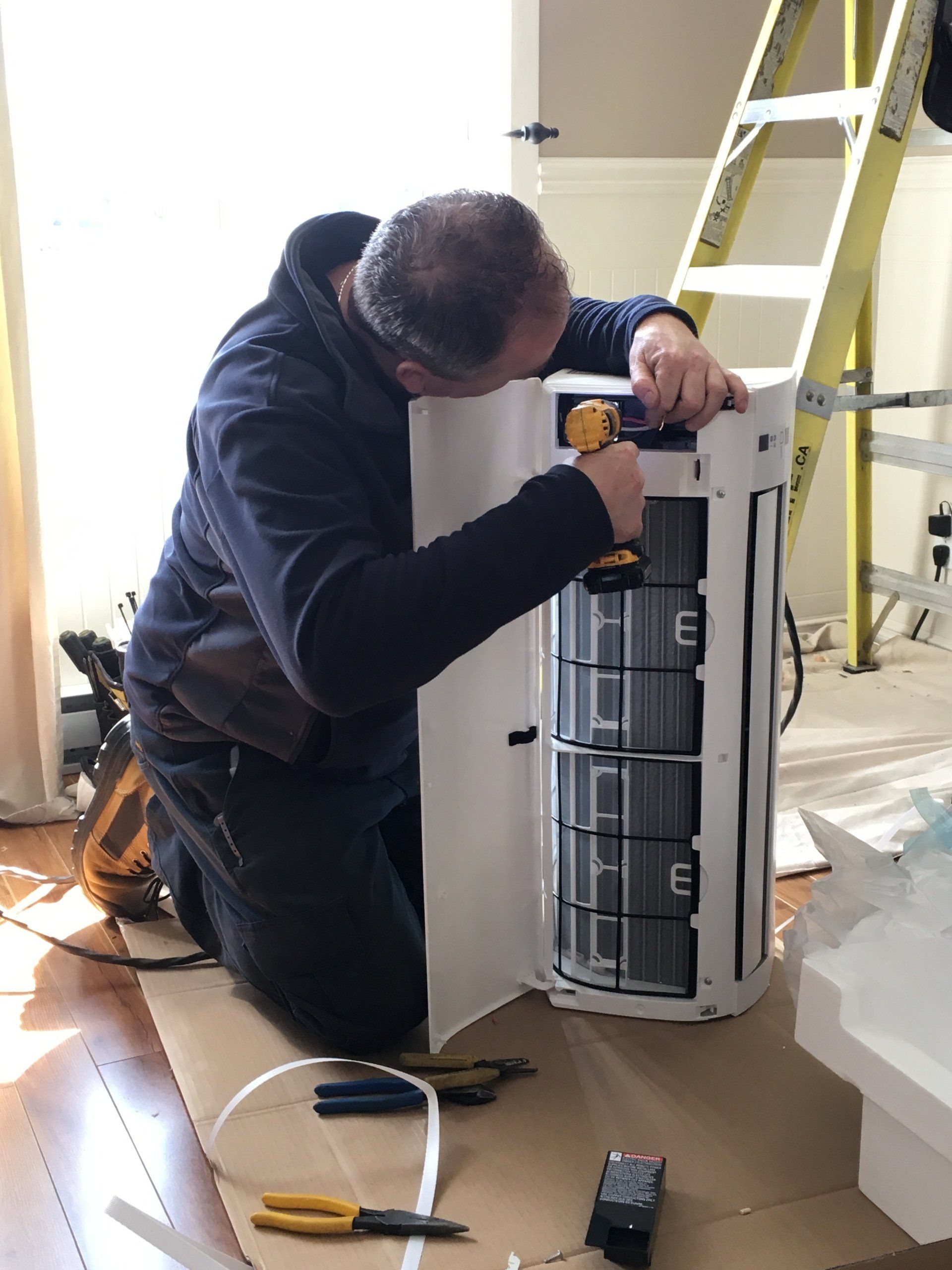 Une personne utilise une perceuse sur un climatiseur blanc. À l'intérieur de l'appareil se trouvent des filtres noirs.