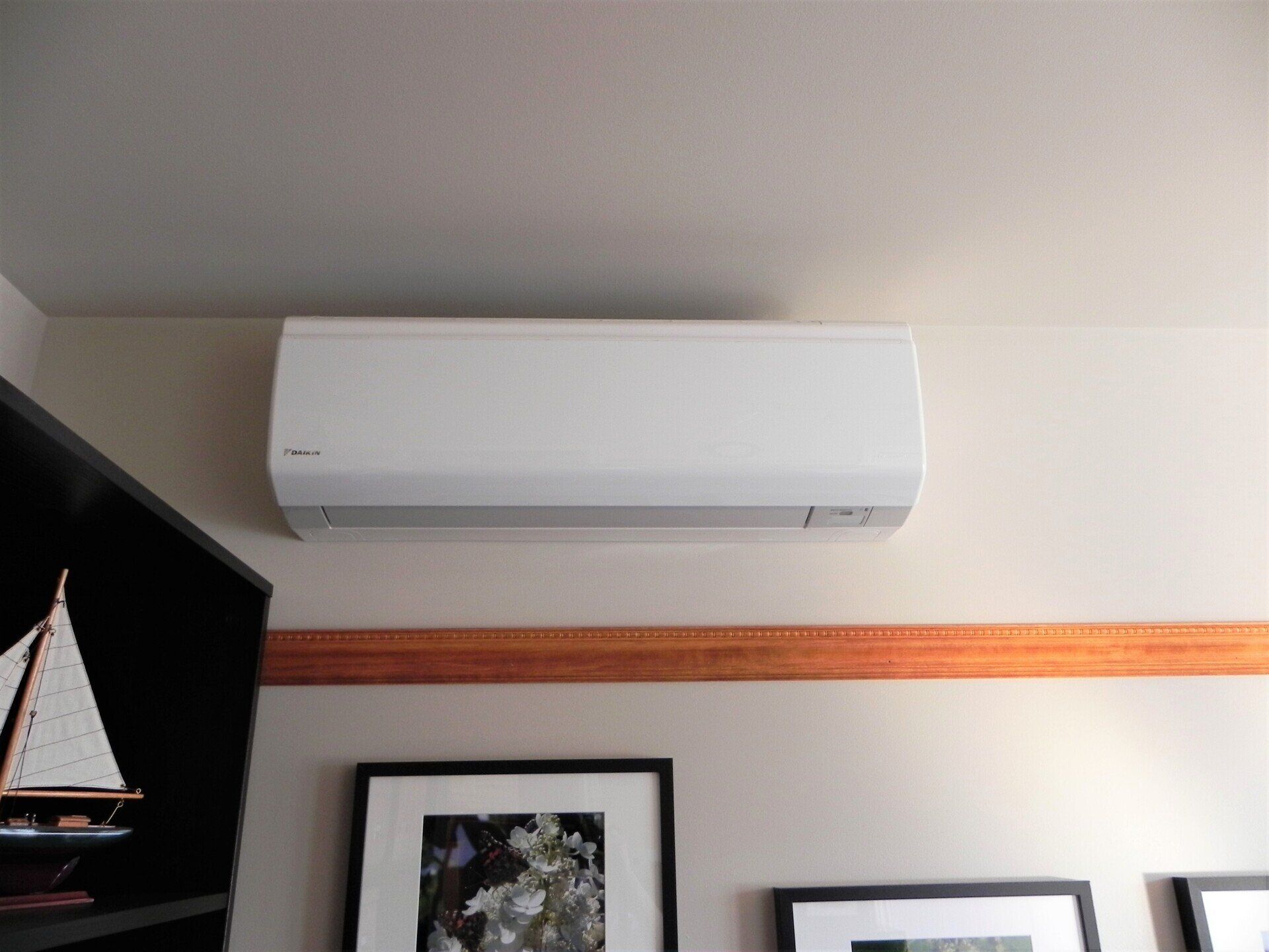 Climatiseur blanc sans conduit, fixé sur un mur beige, au-dessus d'œuvres d'art encadrées et de moulures en bois.