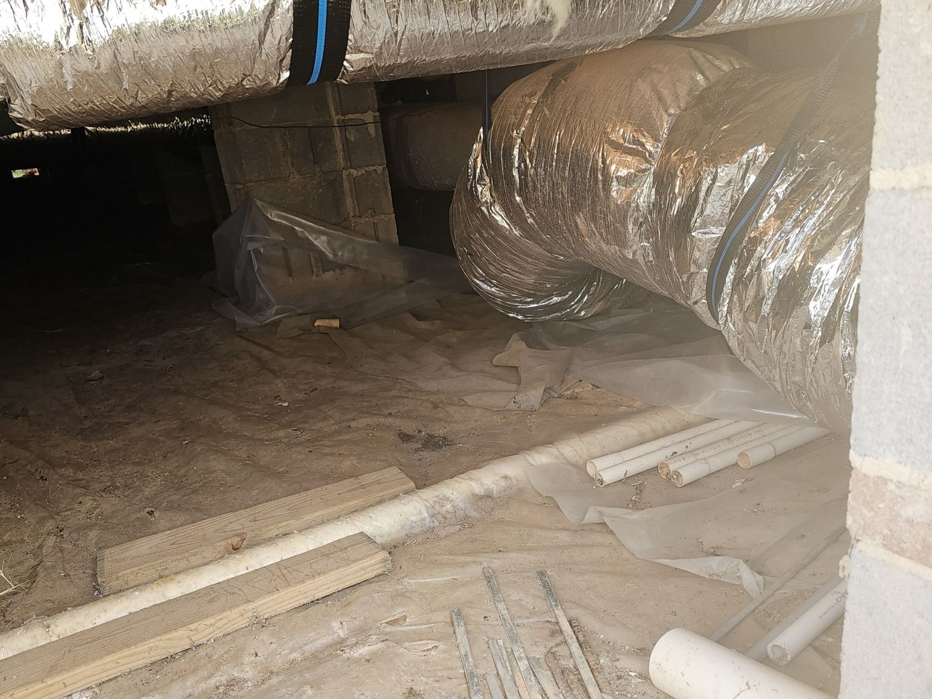View of a crawl space under a house. Ductwork is wrapped in silver insulation. Plastic sheeting and scattered debris are on the dirt floor.