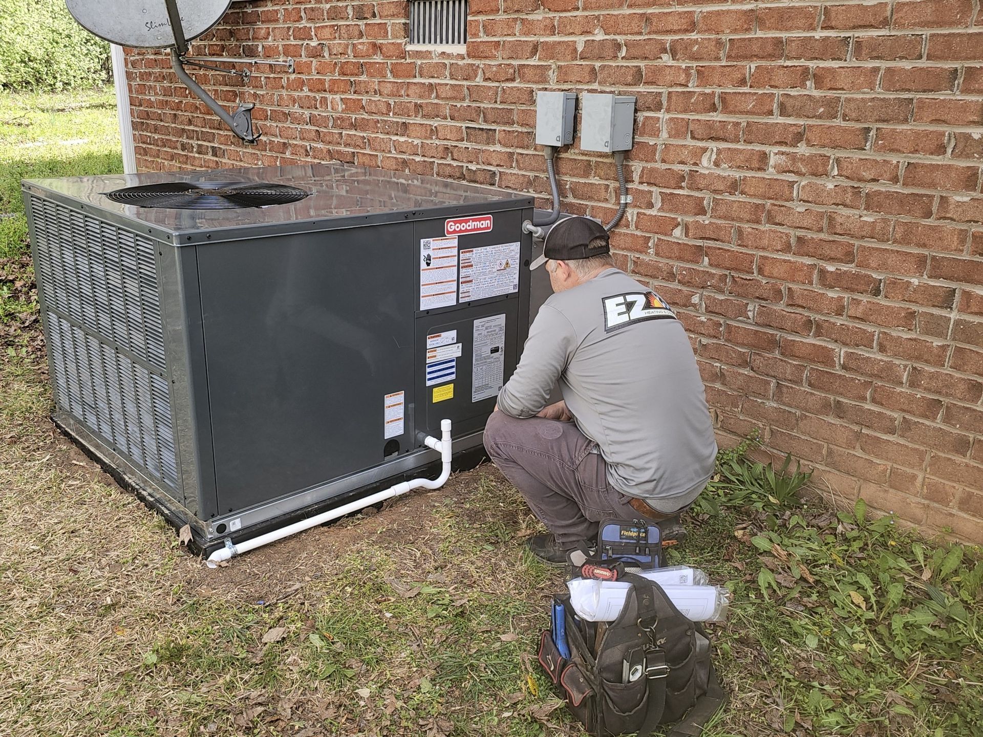 A technician crouches near a new air conditioning unit installed outside a brick building, inspecting it.