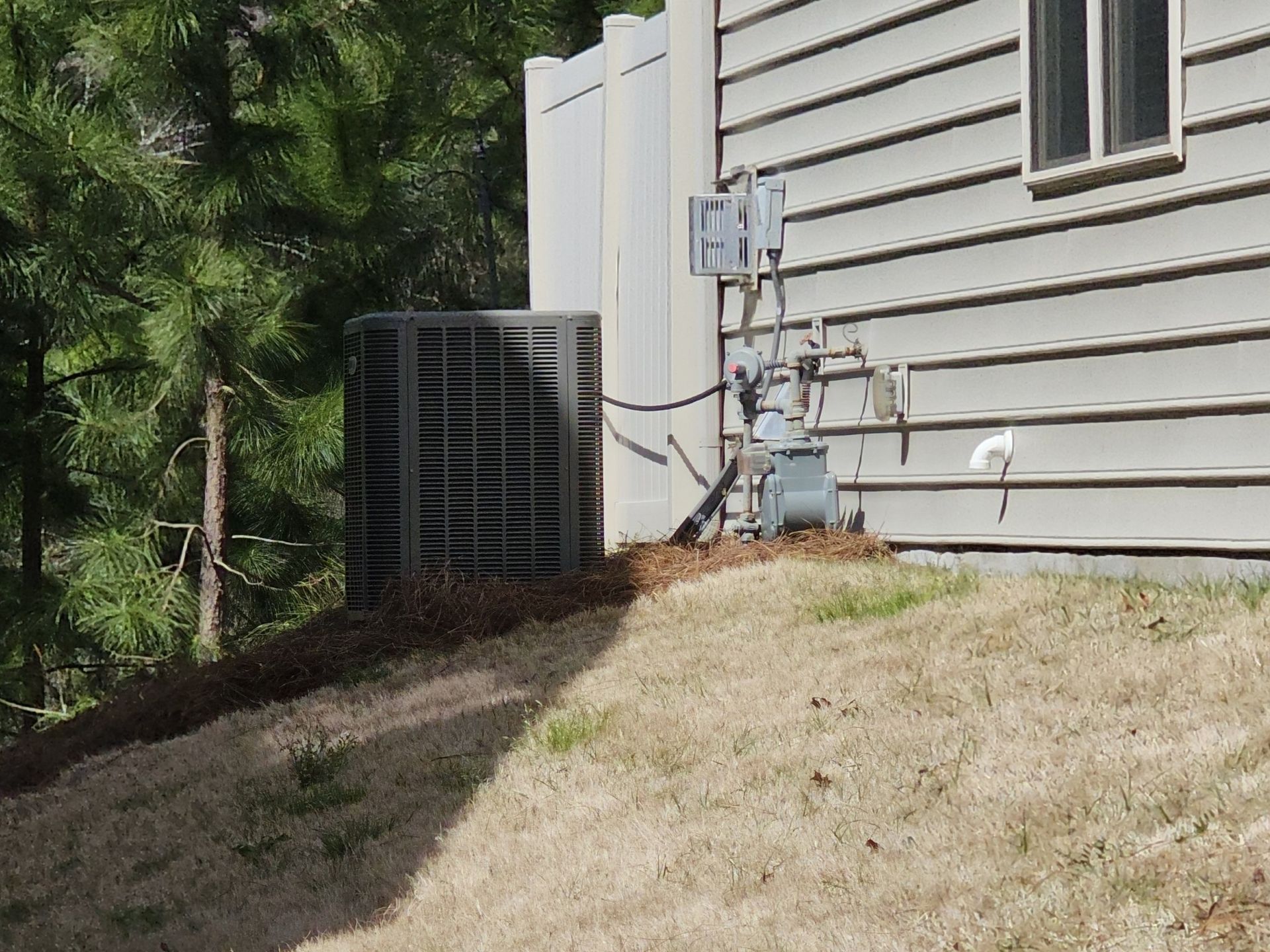Air conditioner unit next to a house with siding, gas meter, and electrical components. The unit is on a slight hill with dry grass.