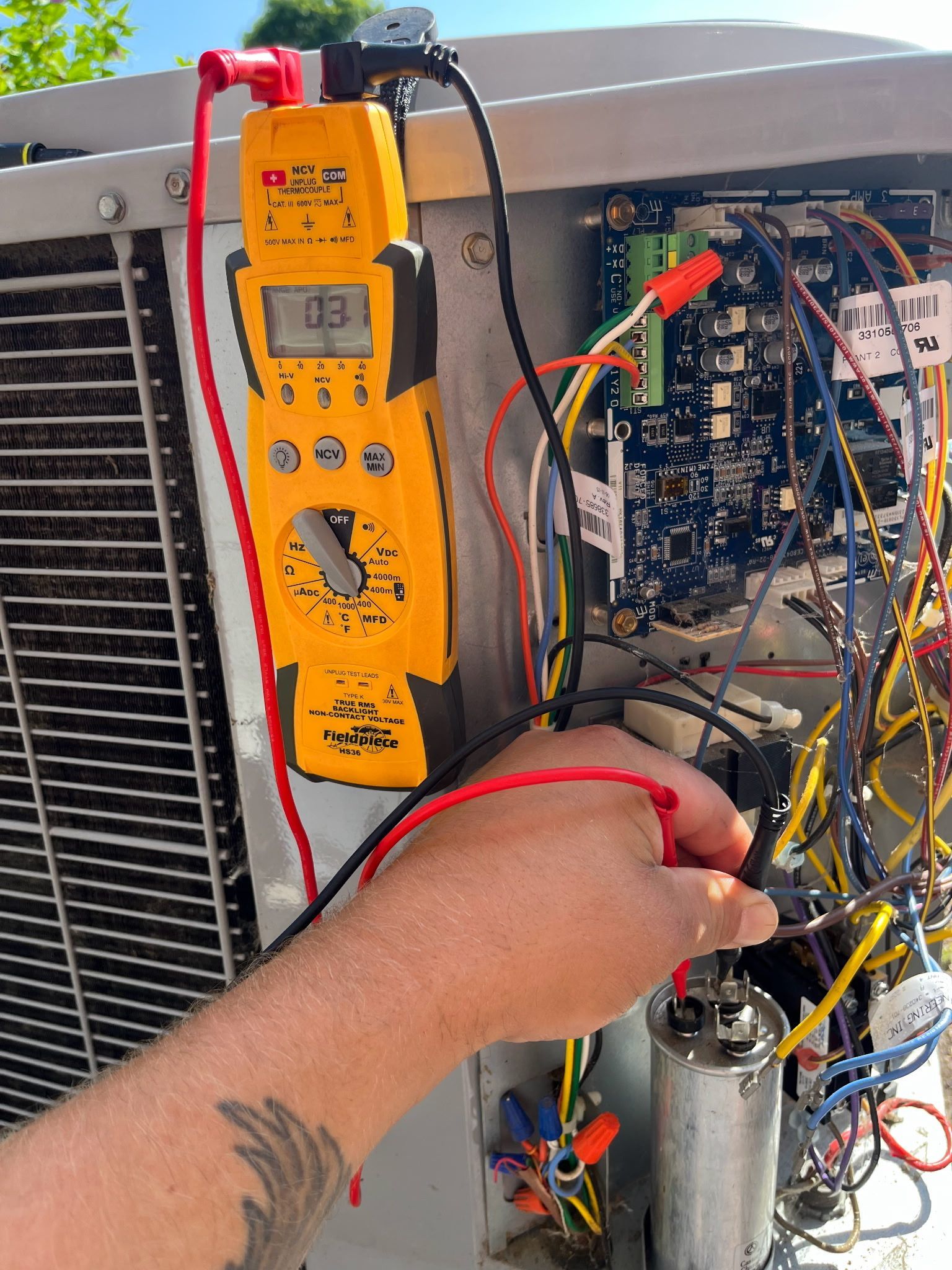 A technician uses a multimeter on the electrical components of an outdoor AC unit. Yellow meter reads 0.3, various wires and circuit board are visible.