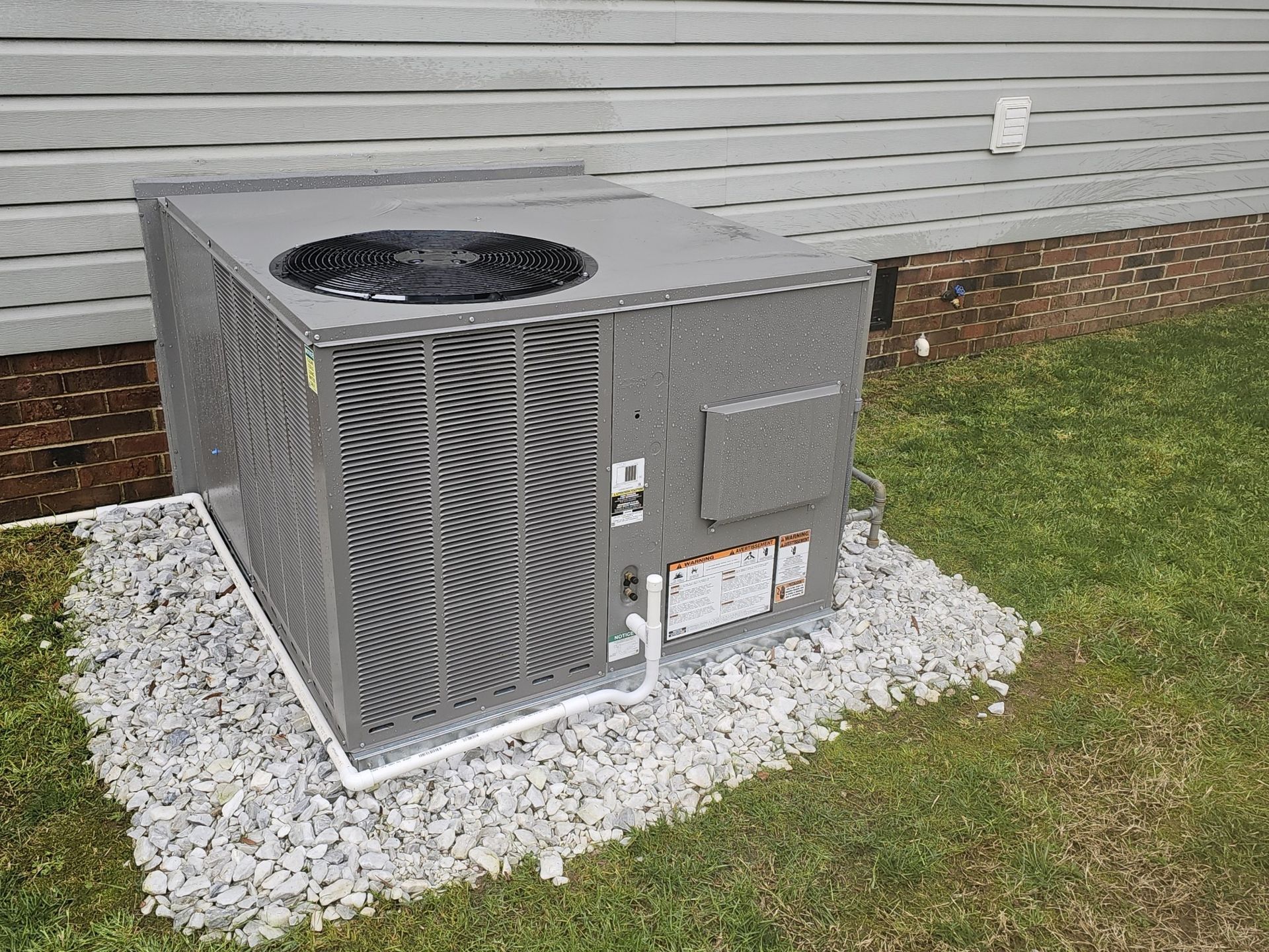 Exterior HVAC unit on a bed of gravel next to a house with gray siding.  A white pipe runs along the base of the unit.