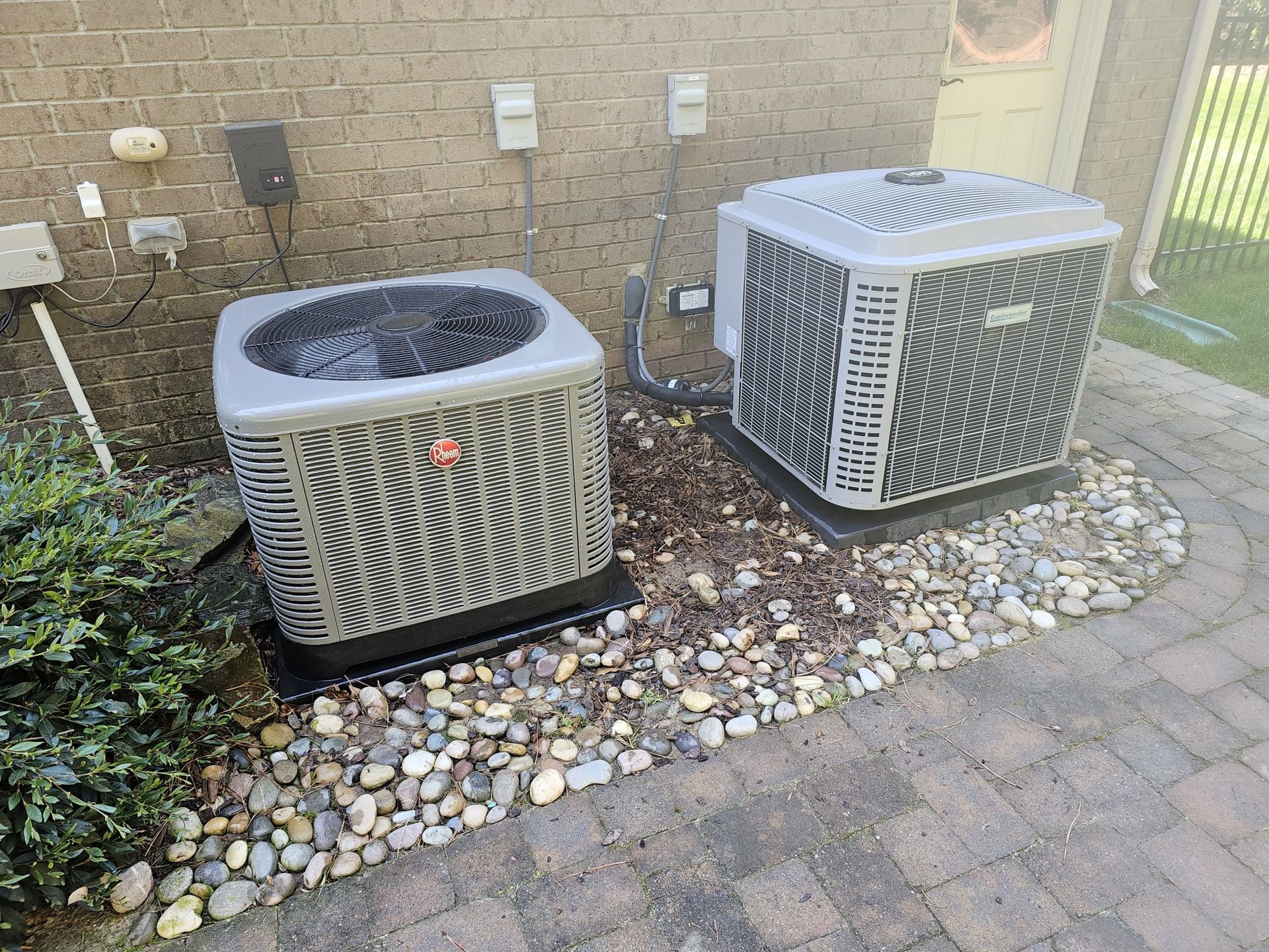Two gray air conditioning units sit on a brick patio next to a brick wall. Rocks and foliage surround them.