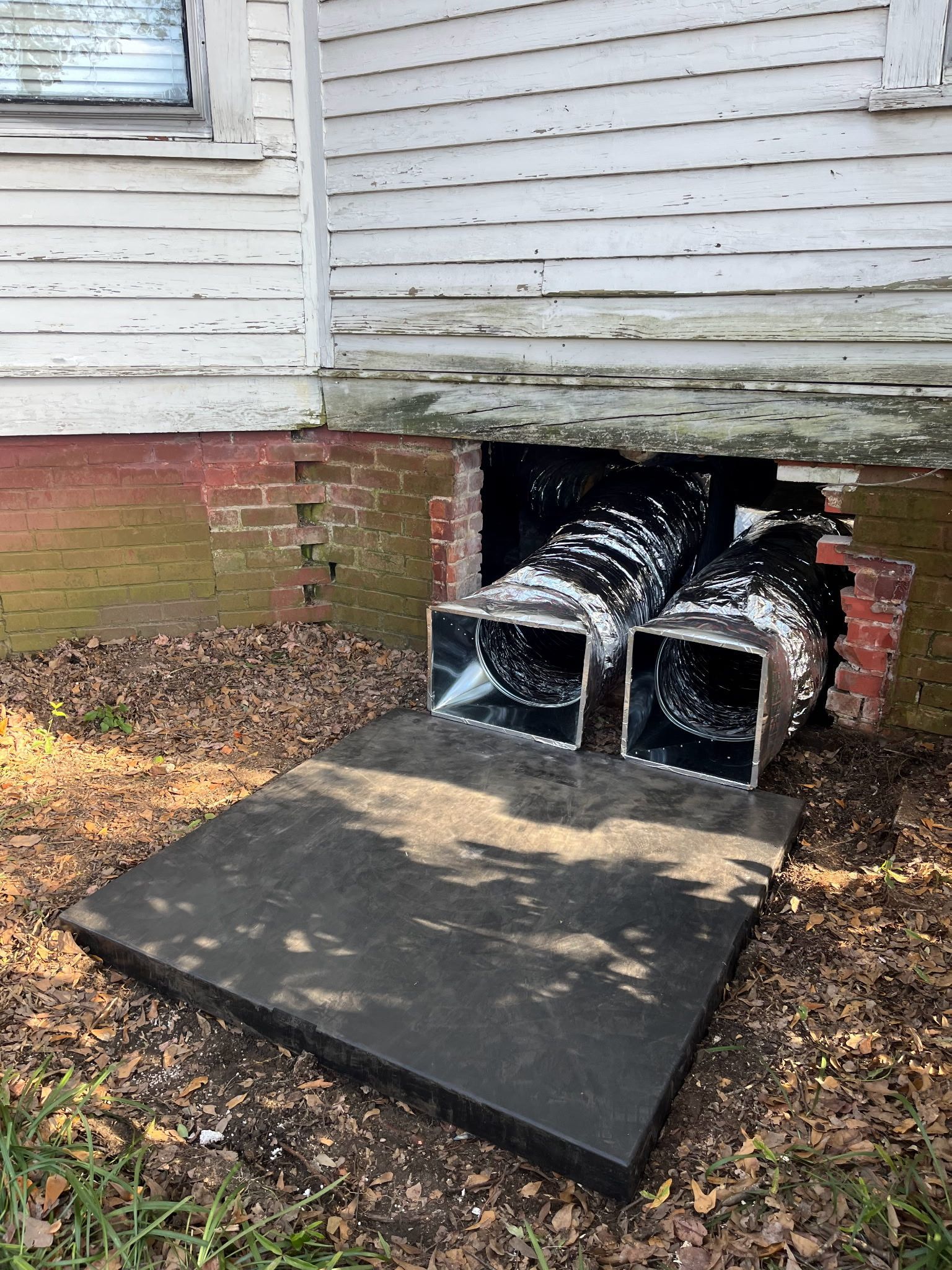 Exterior of a weathered building with two vents exiting the foundation above a dark, square slab. The building is white with brick and peeling paint.
