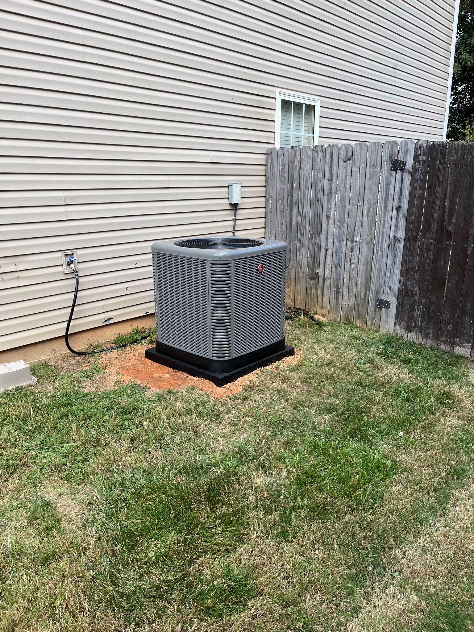 An air conditioning unit sits in a grassy yard next to a wooden fence and beige siding.