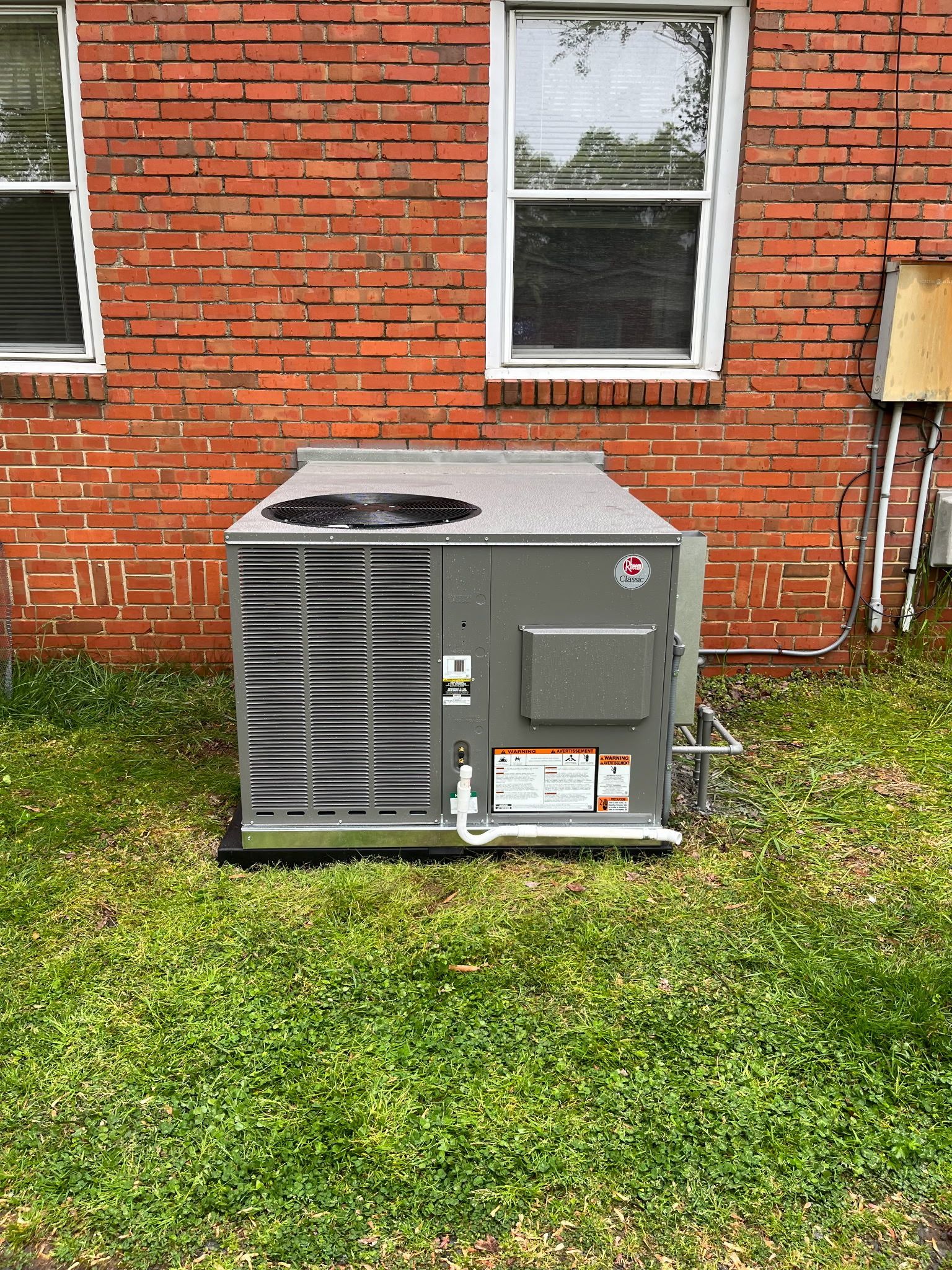 A gray air conditioning unit sits on a black base against a red brick wall, grass surrounds it.
