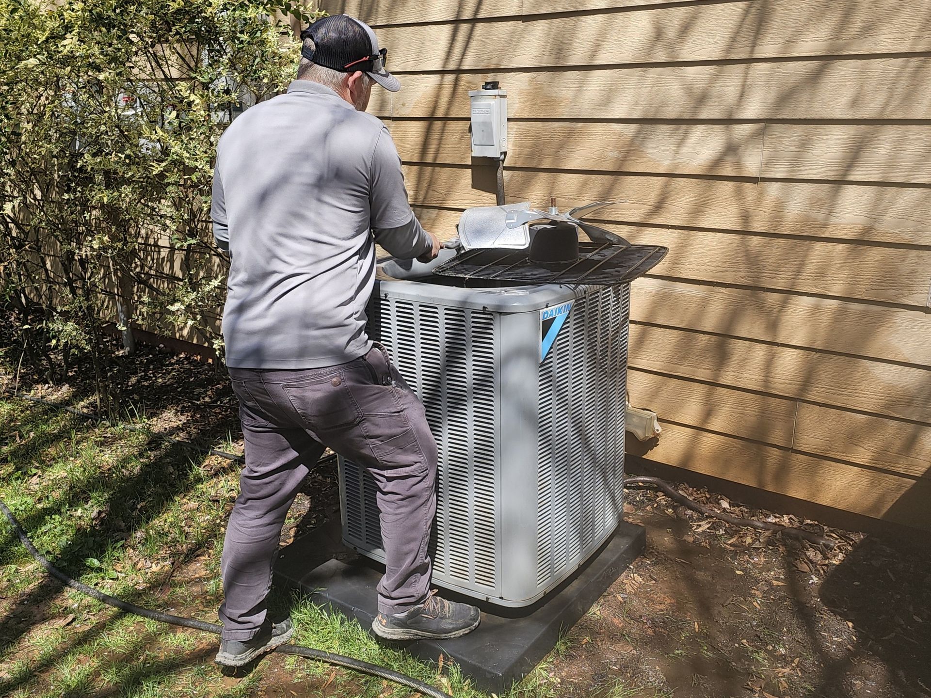 A person in gray clothing inspects an outdoor air conditioning unit next to a wooden building.