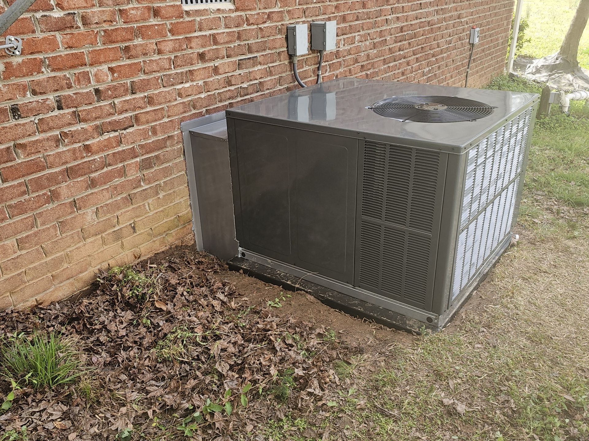A dark-colored air conditioning unit sits next to a red brick wall, on a bed of leaves and grass.