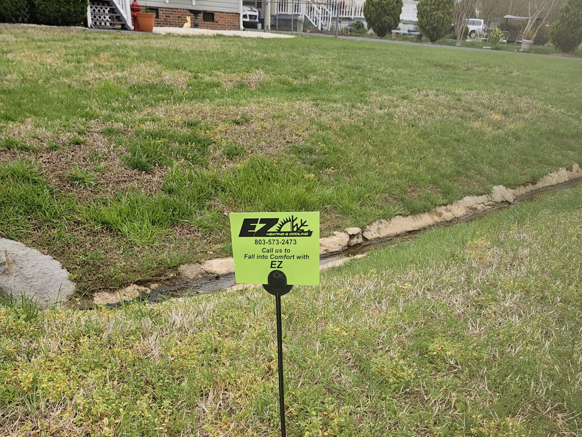 A sign that reads EZ Lawn Protection is in a grassy area near a small drainage ditch. In the background are houses and other vegetation.
