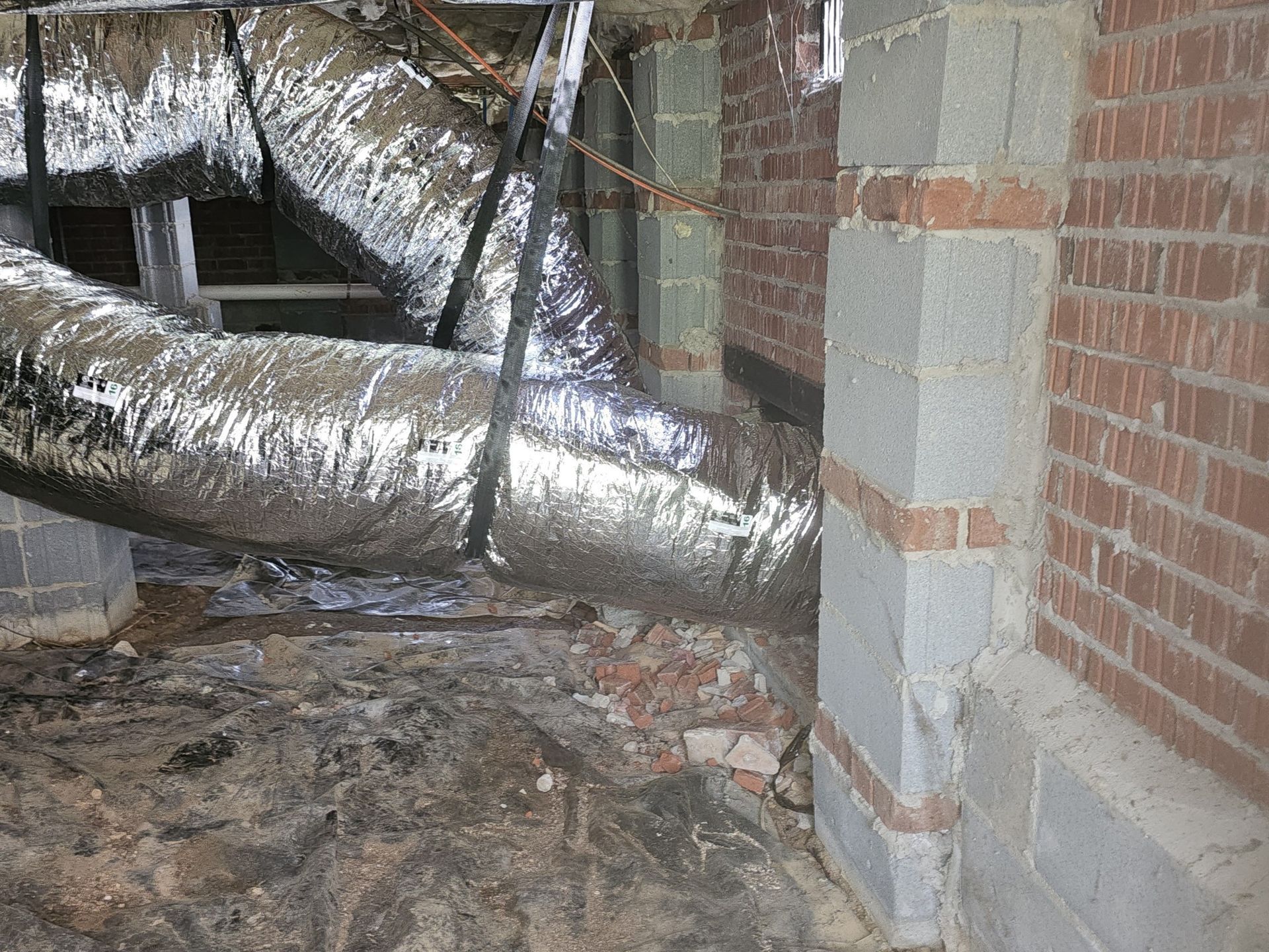 Air ducts snaking through a crawl space with brick and cinder block supports and a damaged brick wall.