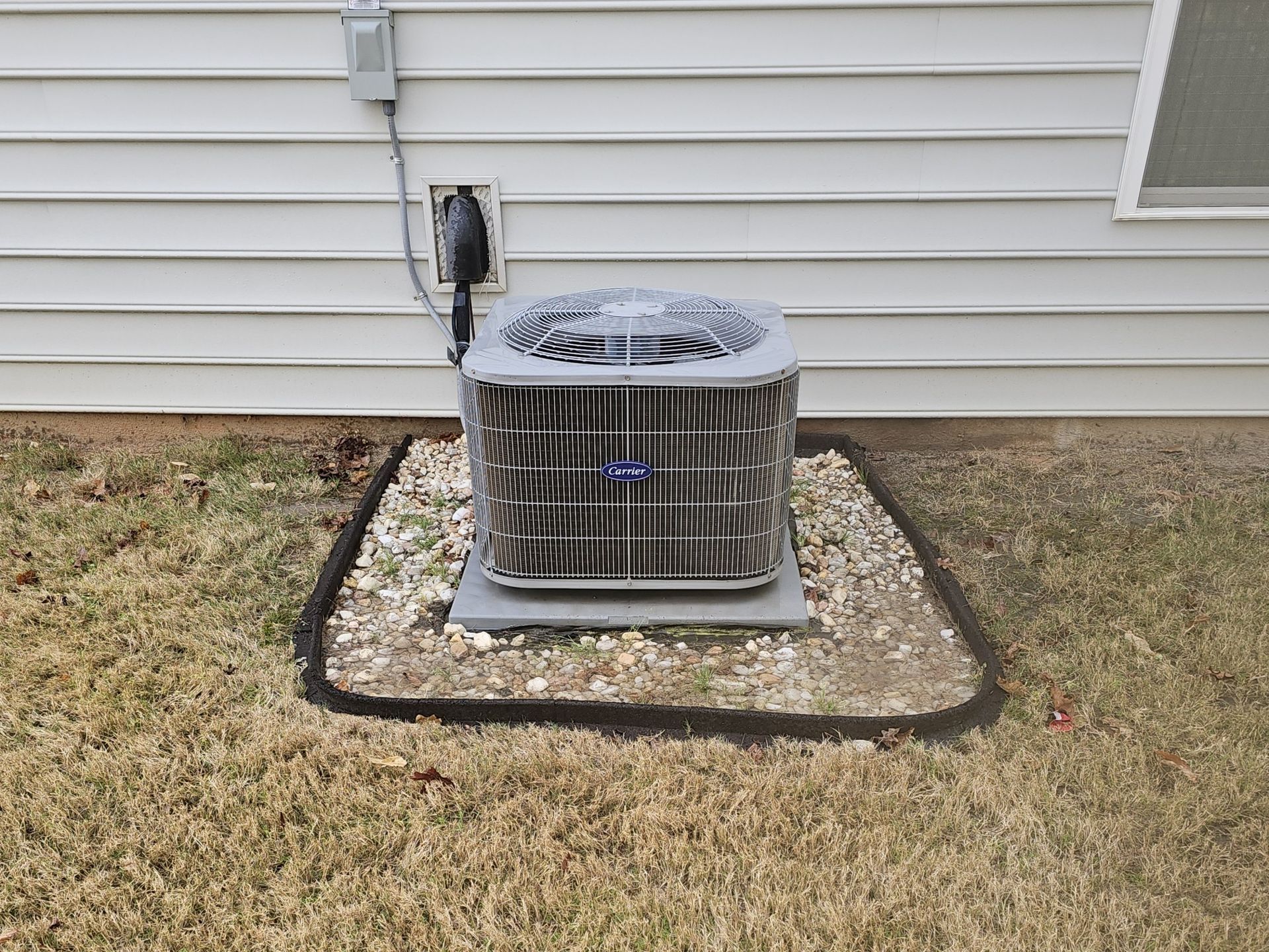 An air conditioning unit sits on a gravel bed in a yard next to a light-colored house.