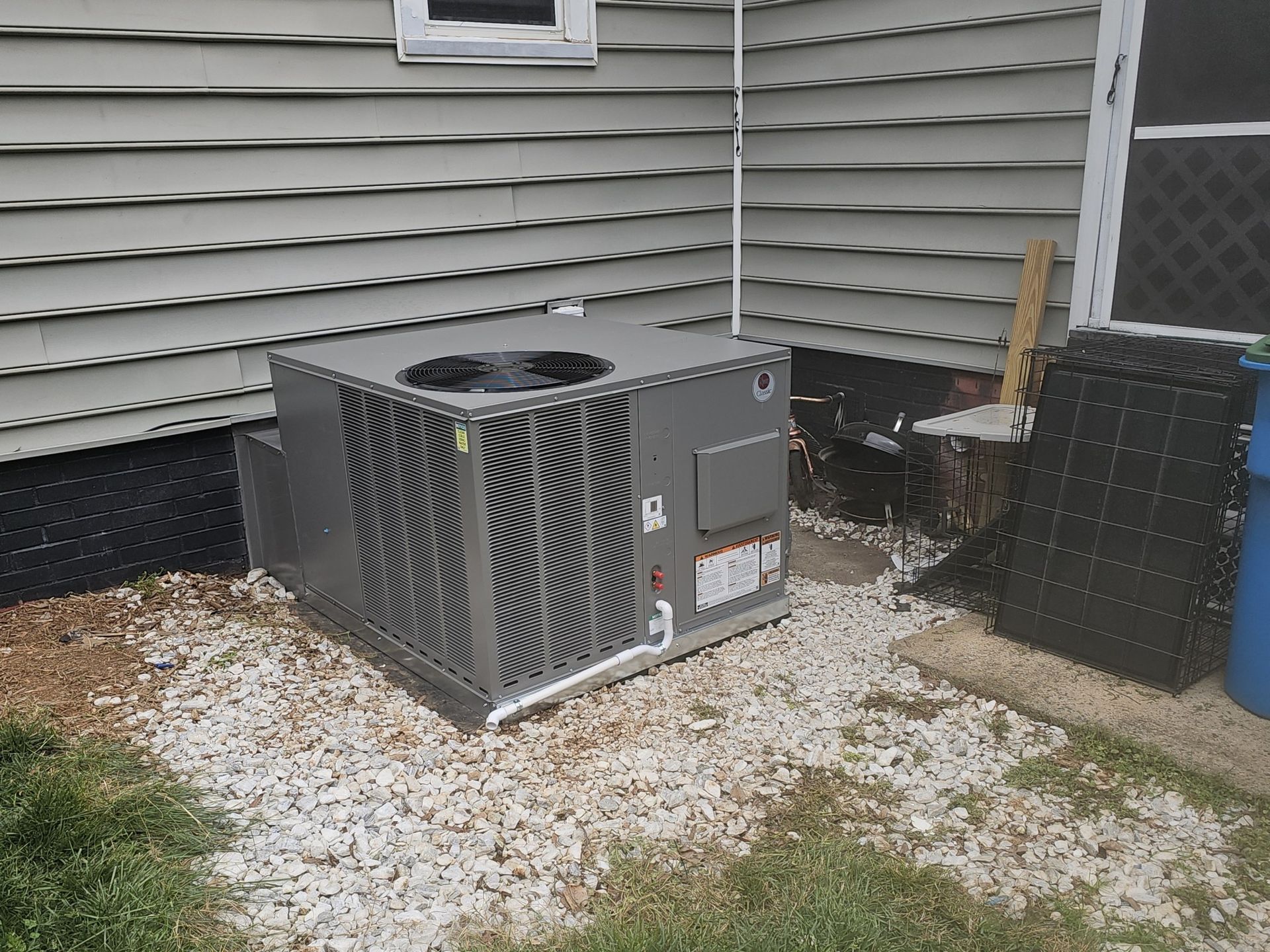 An air conditioning unit, gray in color, sits on gravel beside a house with gray siding.