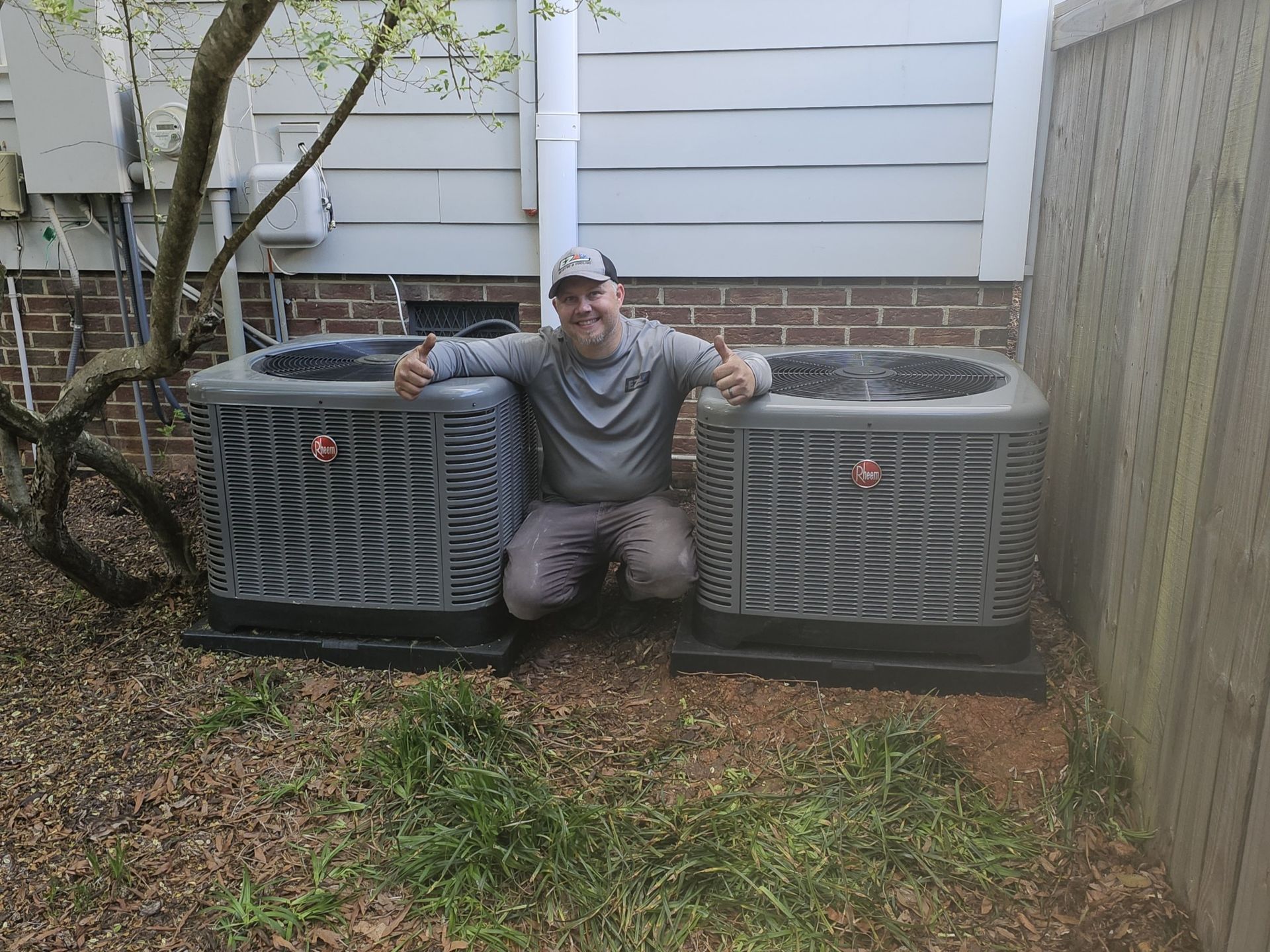 Man kneels between two new air conditioning units, giving thumbs up. He's outside in a backyard with a tree and fence.