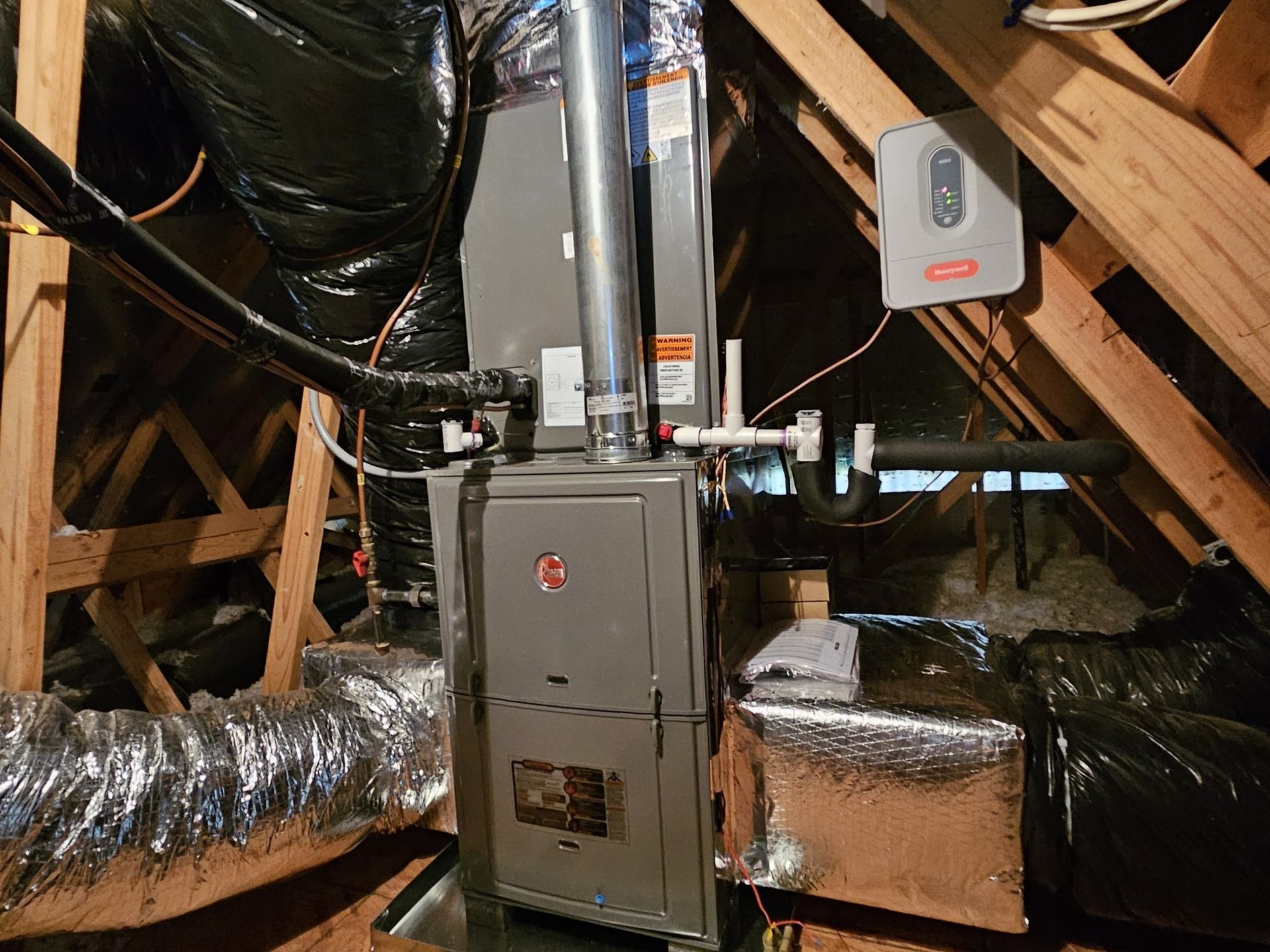 Furnace installation in an attic. A gray furnace is surrounded by ductwork, insulation, and wooden beams.