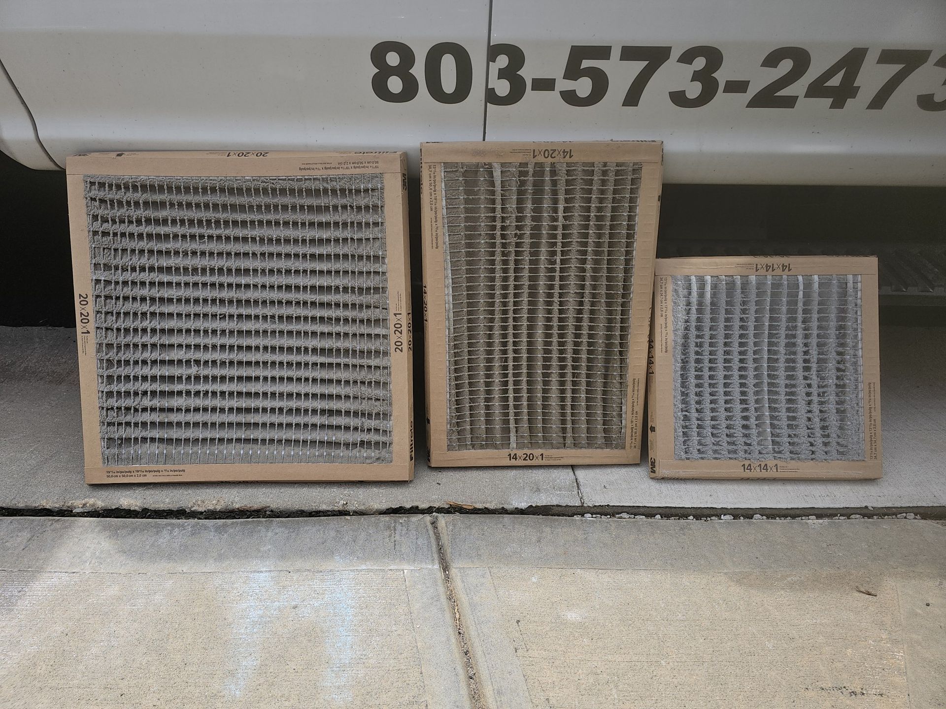 Three used air filters of varying sizes sit on a sidewalk, against a vehicle with a phone number.