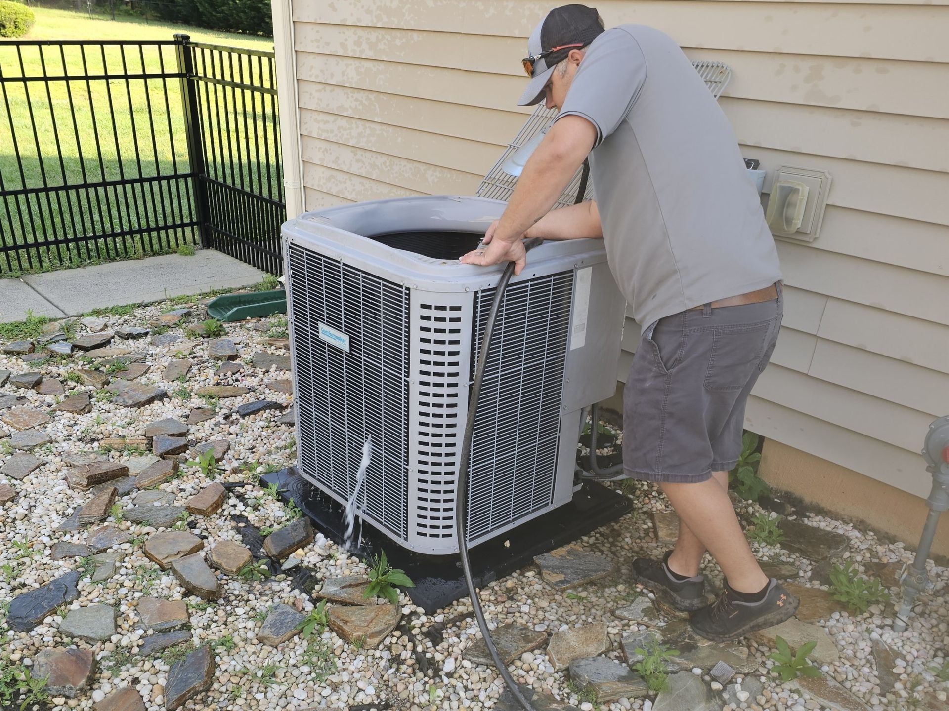 Man in shorts and a t-shirt working on an air conditioning unit outside a house.