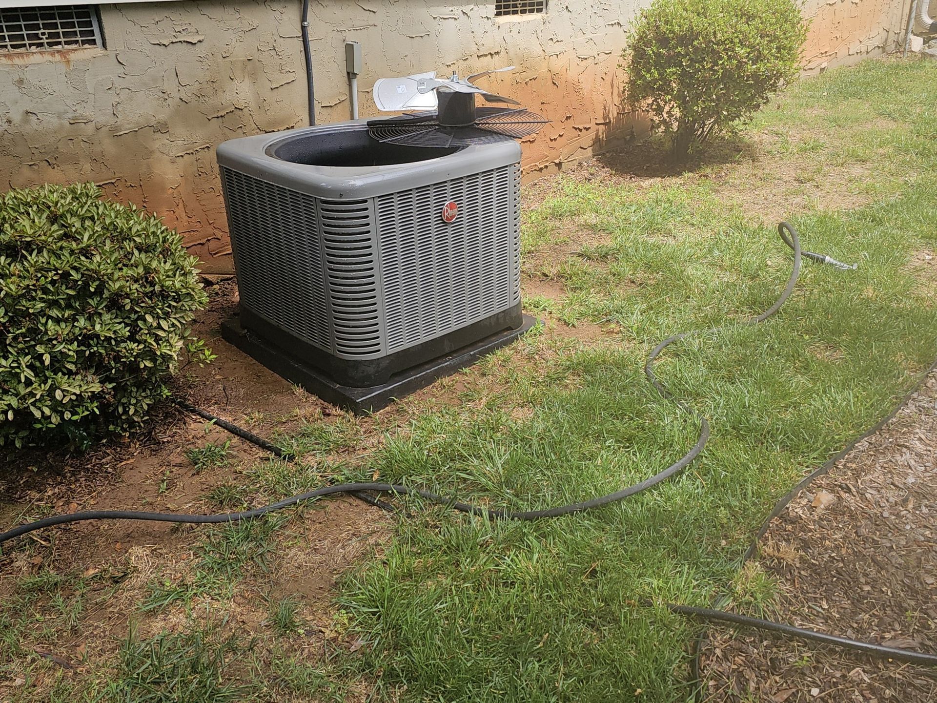 An outdoor air conditioning unit on a black base, surrounded by green grass and a small bush, against a light-colored building.