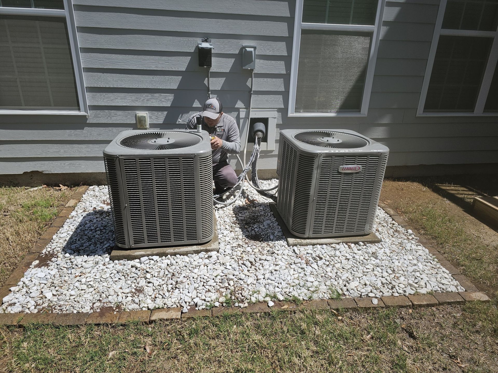 Two air conditioning units on a gravel bed, with a person working on the electrical connections against a light gray house wall.