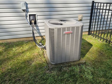 An air conditioning unit sits on a concrete pad outside a light-colored house, next to a black fence.