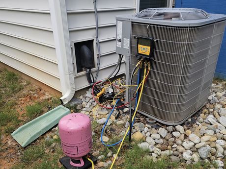 An air conditioning unit being serviced outside a house. Pink refrigerant tank, yellow and blue hoses, and diagnostic equipment are connected.