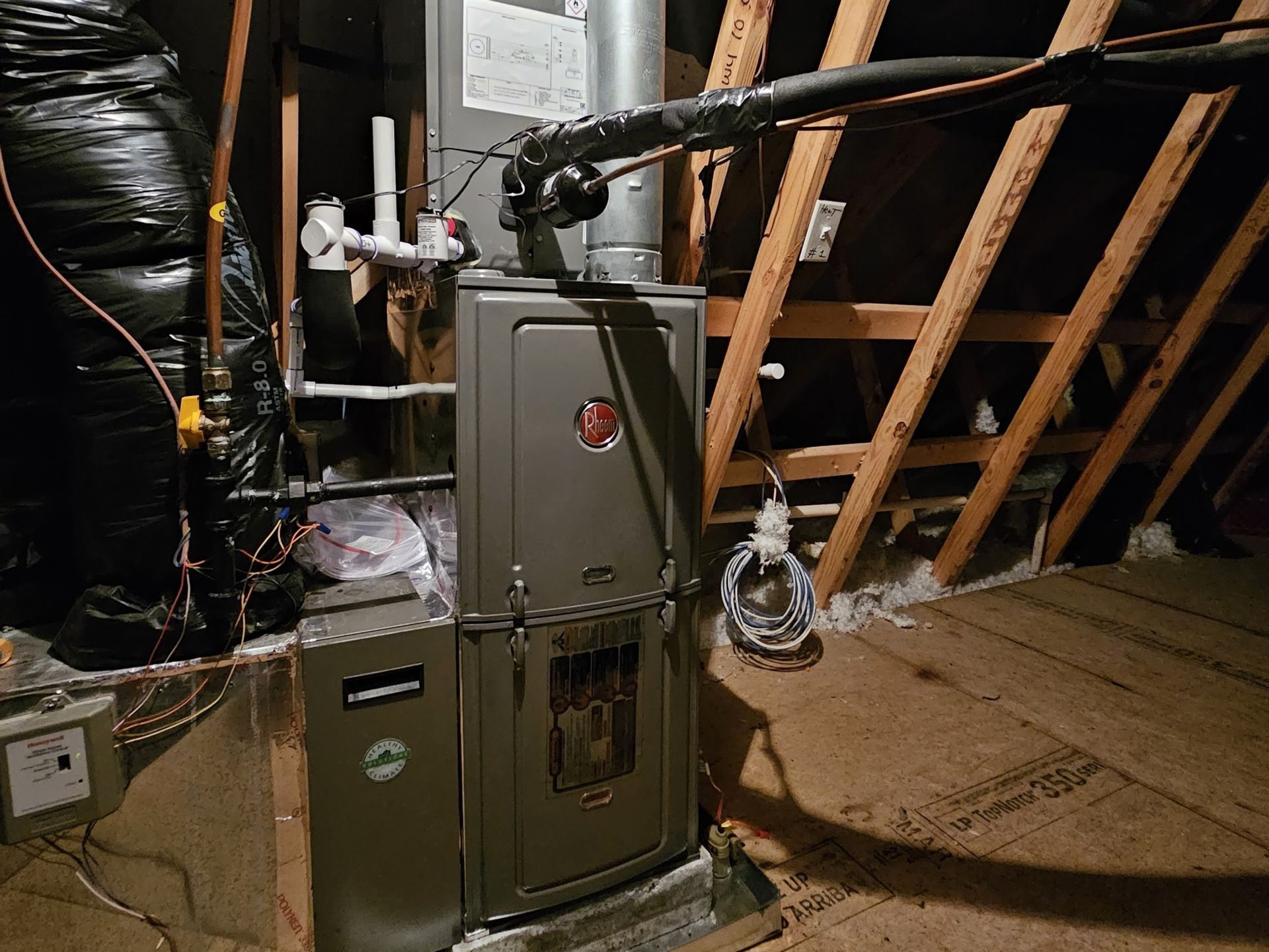 Furnace installation in an attic with exposed wooden beams. The gray furnace is surrounded by insulation and ductwork.