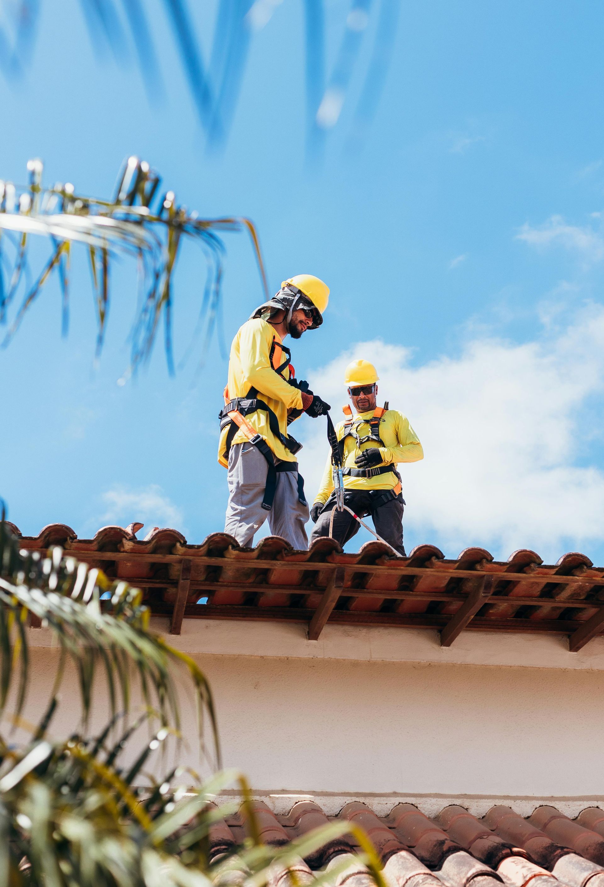 Two construction workers in yellow safety gear on a tiled roof under a blue sky
