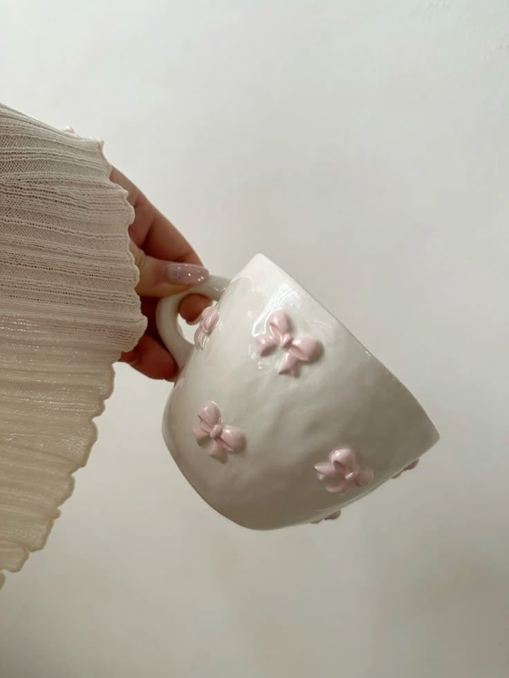 Mano sosteniendo una taza de té blanca decorada con flores rosas sobre un fondo claro.