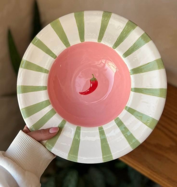 Una mano sostiene un plato blanco con rayas verdes y un cuenco rosa en el centro, adornado con una rodaja de chile rojo.
