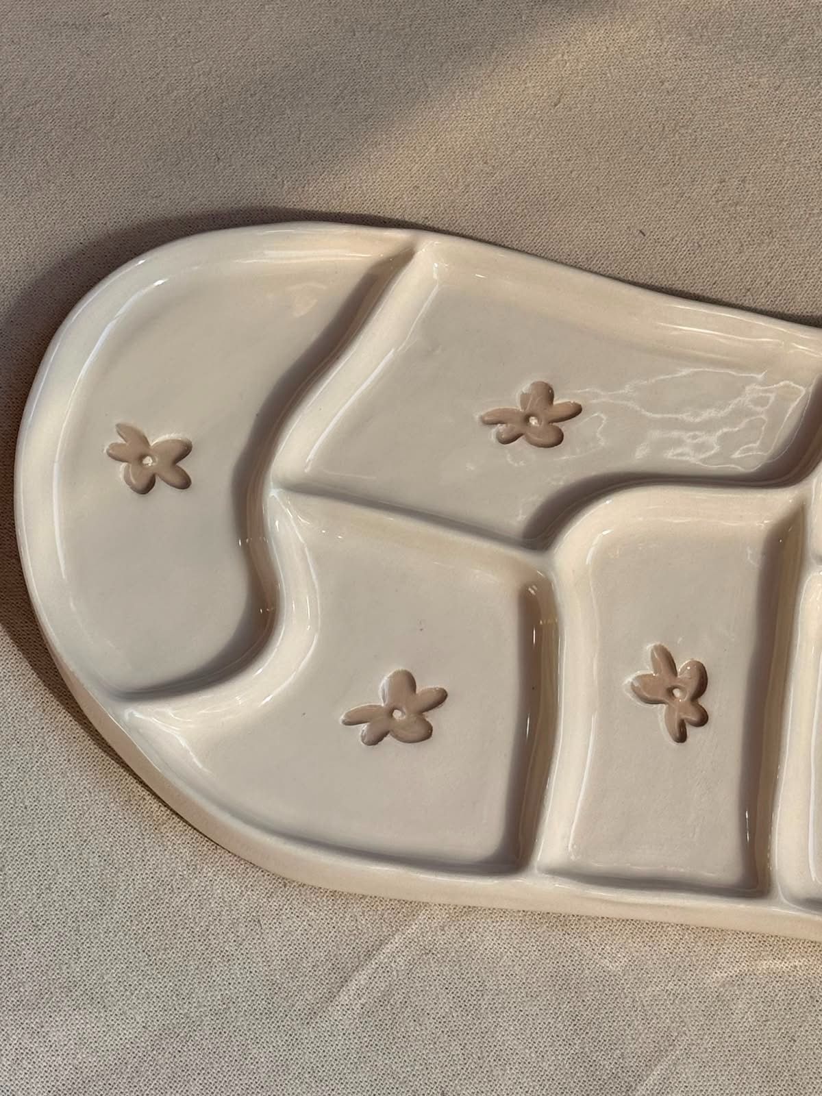 Bandeja de cerámica beige con compartimentos curvos y pequeños detalles en forma de flor sobre una superficie de tela.