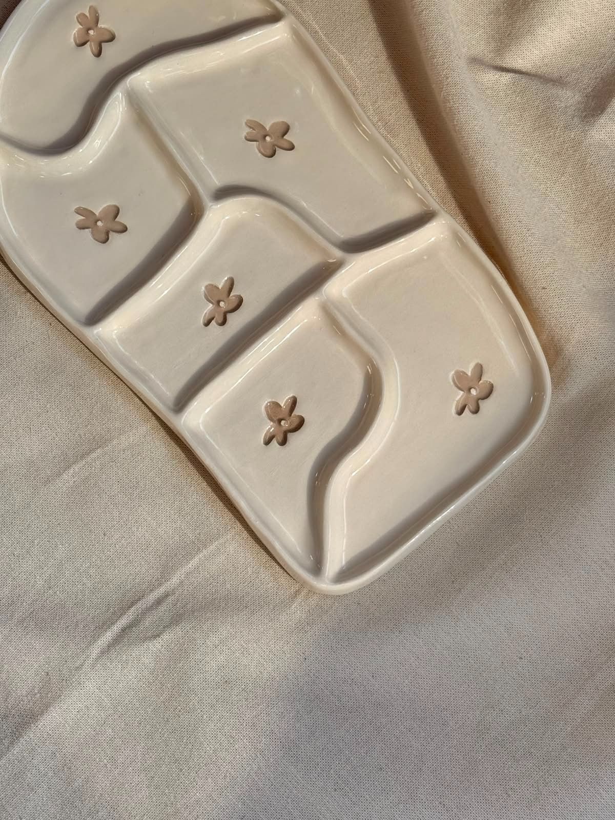 Bandeja de cerámica beige con compartimentos curvos y pequeños detalles en forma de flor sobre una superficie de tela.