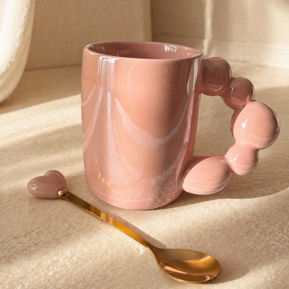 Taza de mármol rosa con asa esculpida y cuchara dorada sobre una superficie iluminada por el sol.
