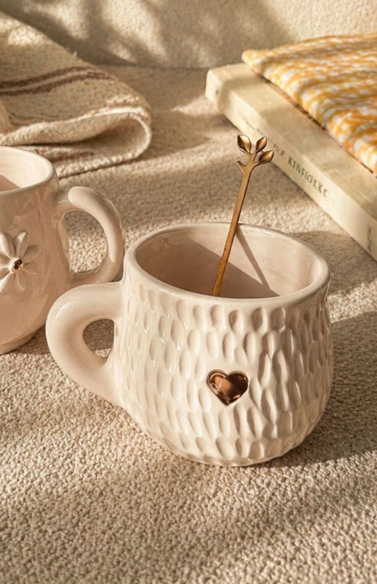 Taza blanca texturizada con detalle de corazón sobre una superficie beige, con una cuchara removiendo el café junto a una tetera y un libro.