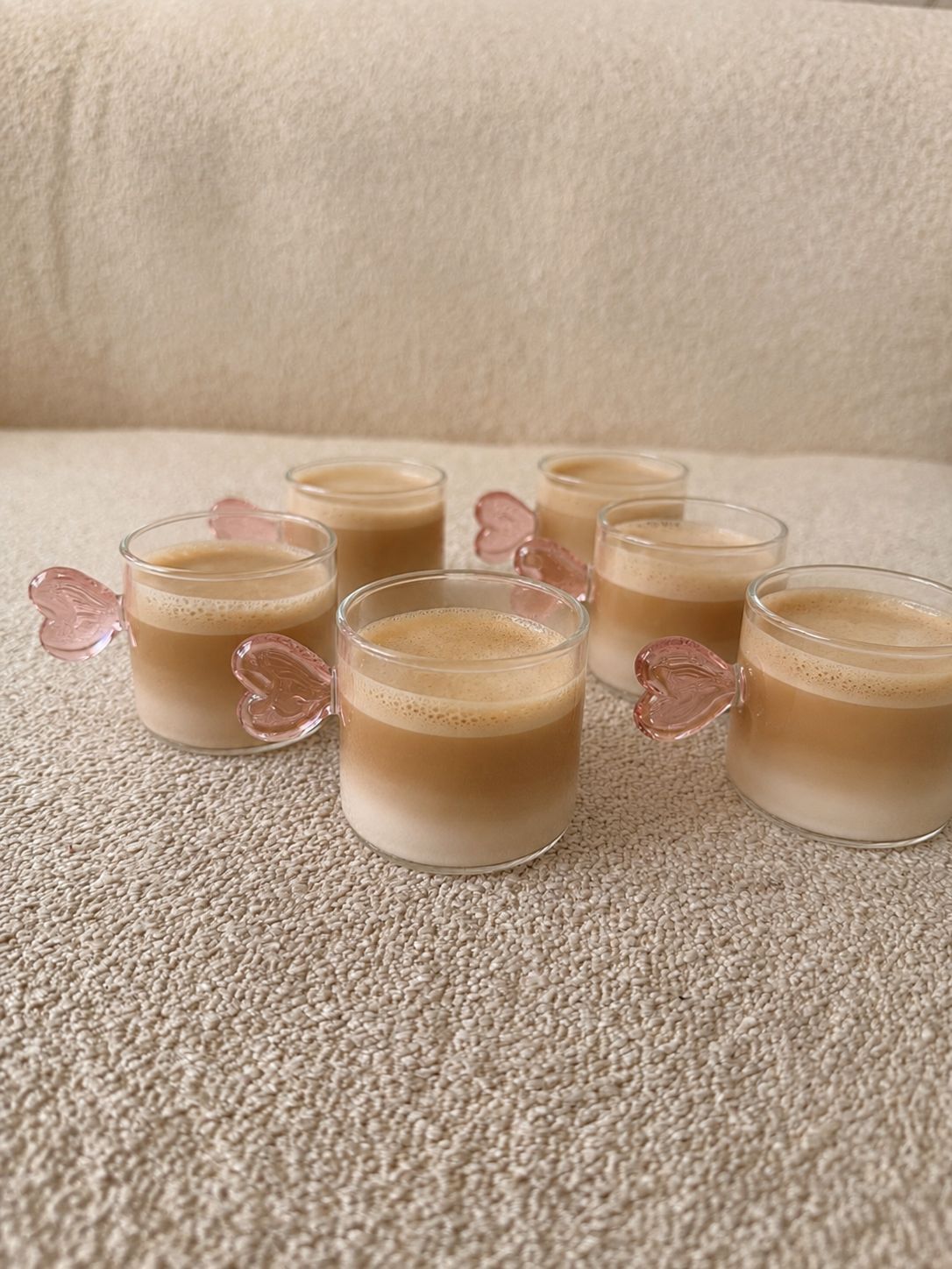Cinco pequeñas velas beige en vasos de cristal transparente sobre una superficie texturizada color crema, con detalles de mariposas rosas.