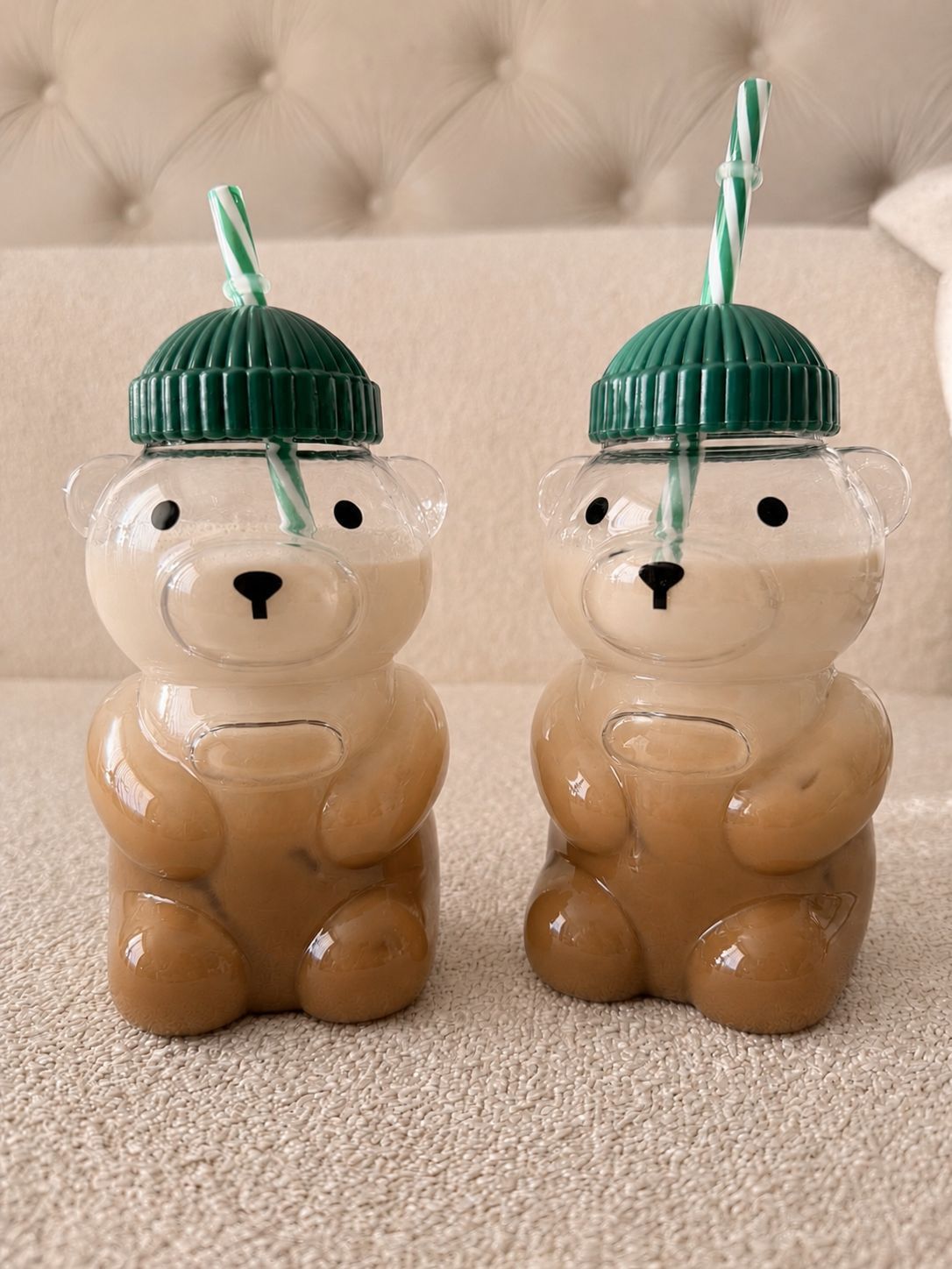 Dos bebidas con forma de oso en vasos transparentes con tapas de pajita verde sobre una alfombra beige.