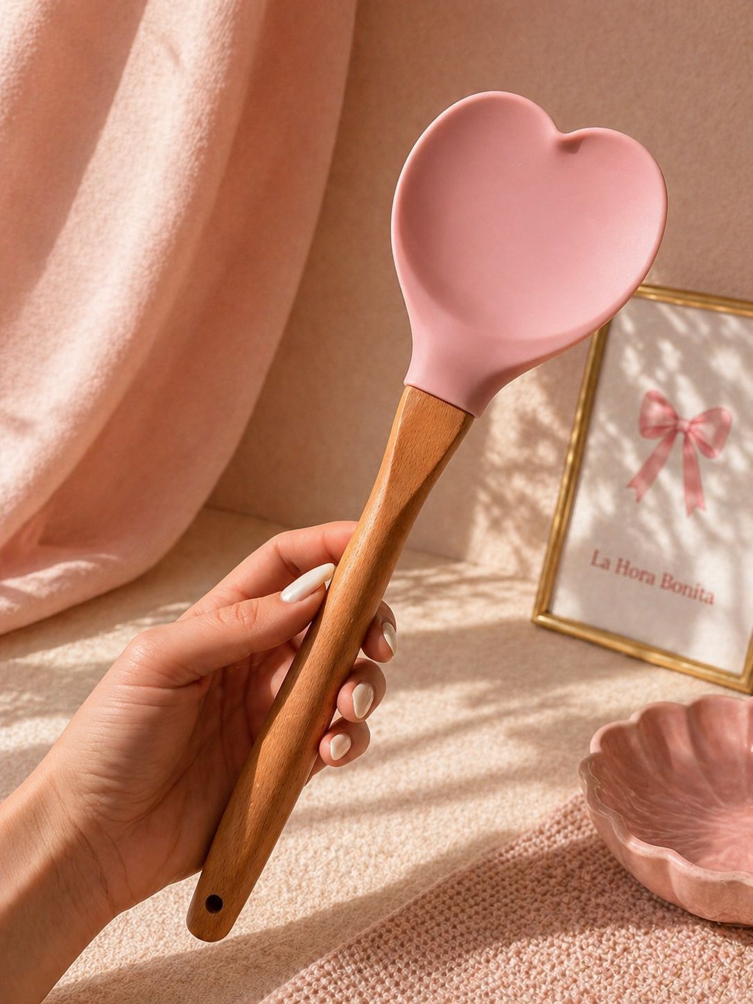 Mano sosteniendo un espejo rosa en forma de corazón con mango de madera sobre un fondo rosa suave.